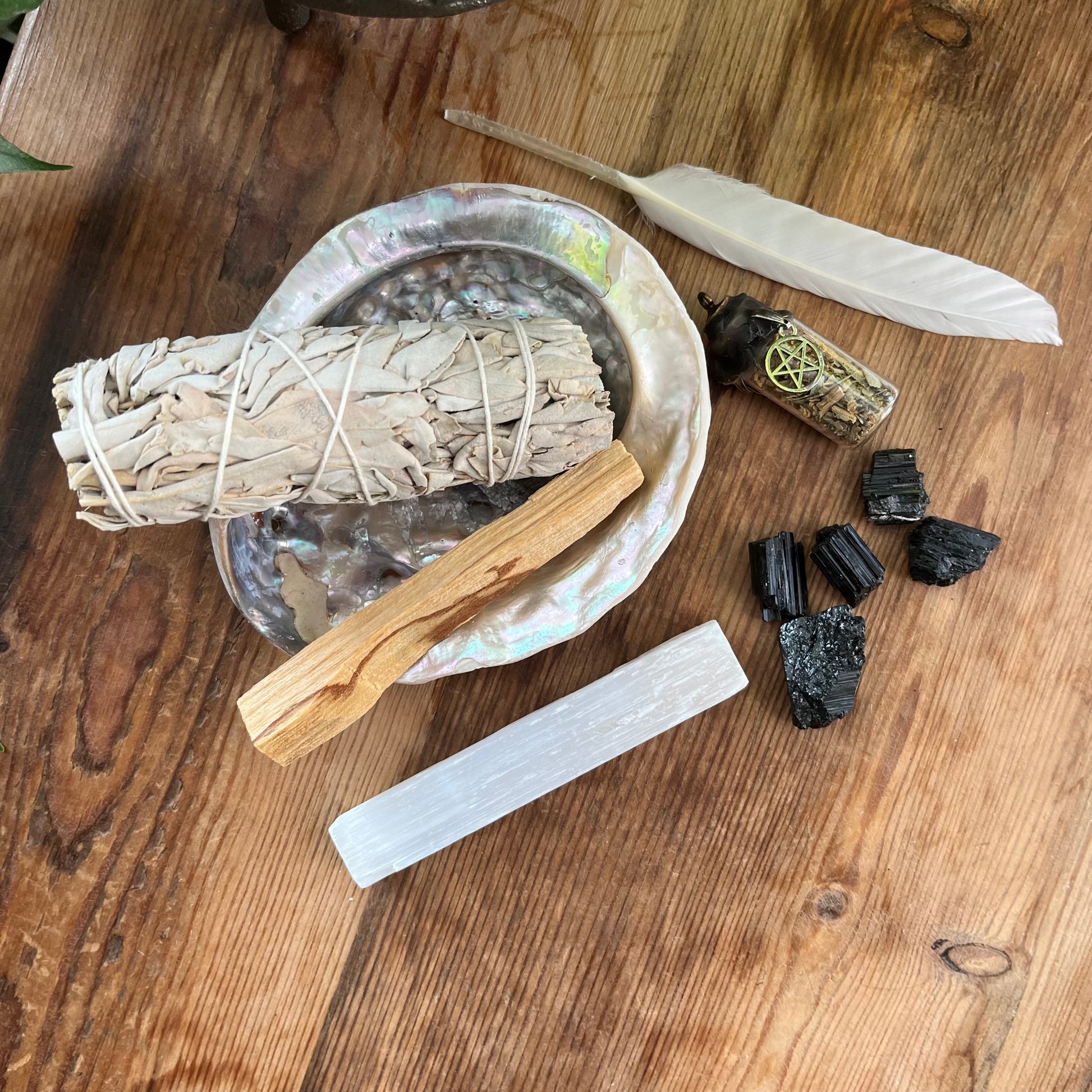 The contents of our 'sacred cleansing ritual gift box' - abalone shell, sage smoke stick, palo santo stick, rough raw selenite stick, 5 rough black tourmaline gemstones and a white goose feather. Photographed on a wooden table with a plant in the background 