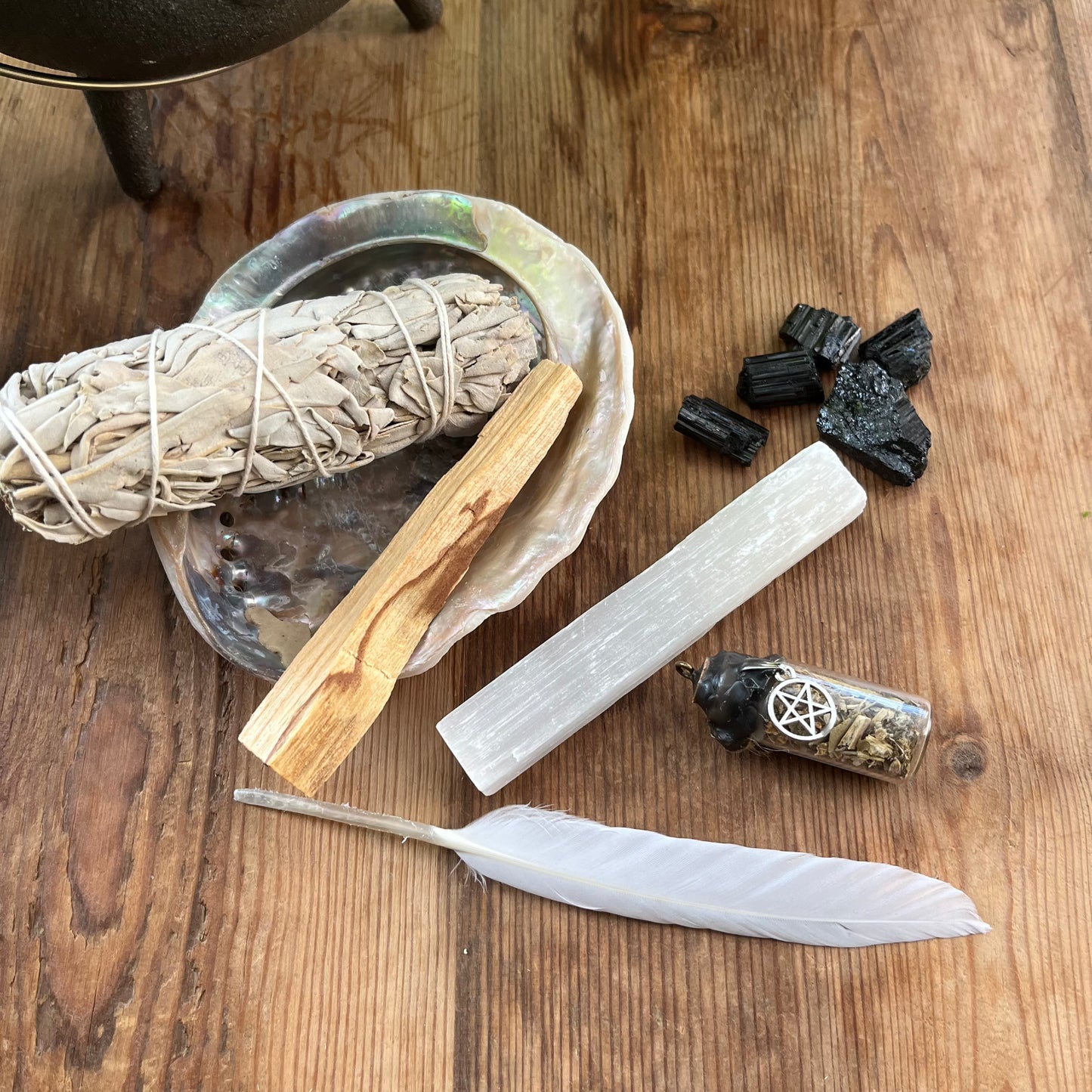 The contents of our 'sacred cleansing ritual gift box' - abalone shell, sage smoke stick, palo santo stick, rough raw selenite stick, 5 rough black tourmaline gemstones and a white goose feather. Photographed on a wooden table with a plant in the background 