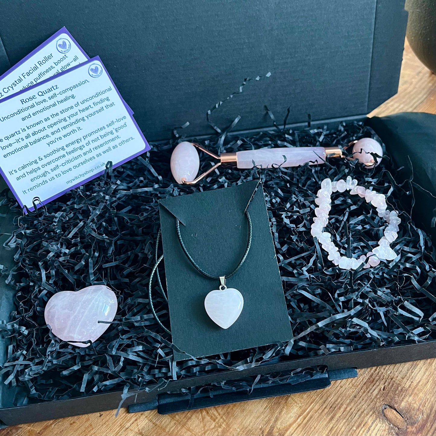 A black gift box with the contents of the Rose Quartz selfcare gift box sat on a wooden table.  rose quartz face roller, rose quartz chip bracelet, a rose quartz heart , and a rose quartz pendent heart necklace. Plus information cards with the properties of rose quartz and benefits of a face roller.  