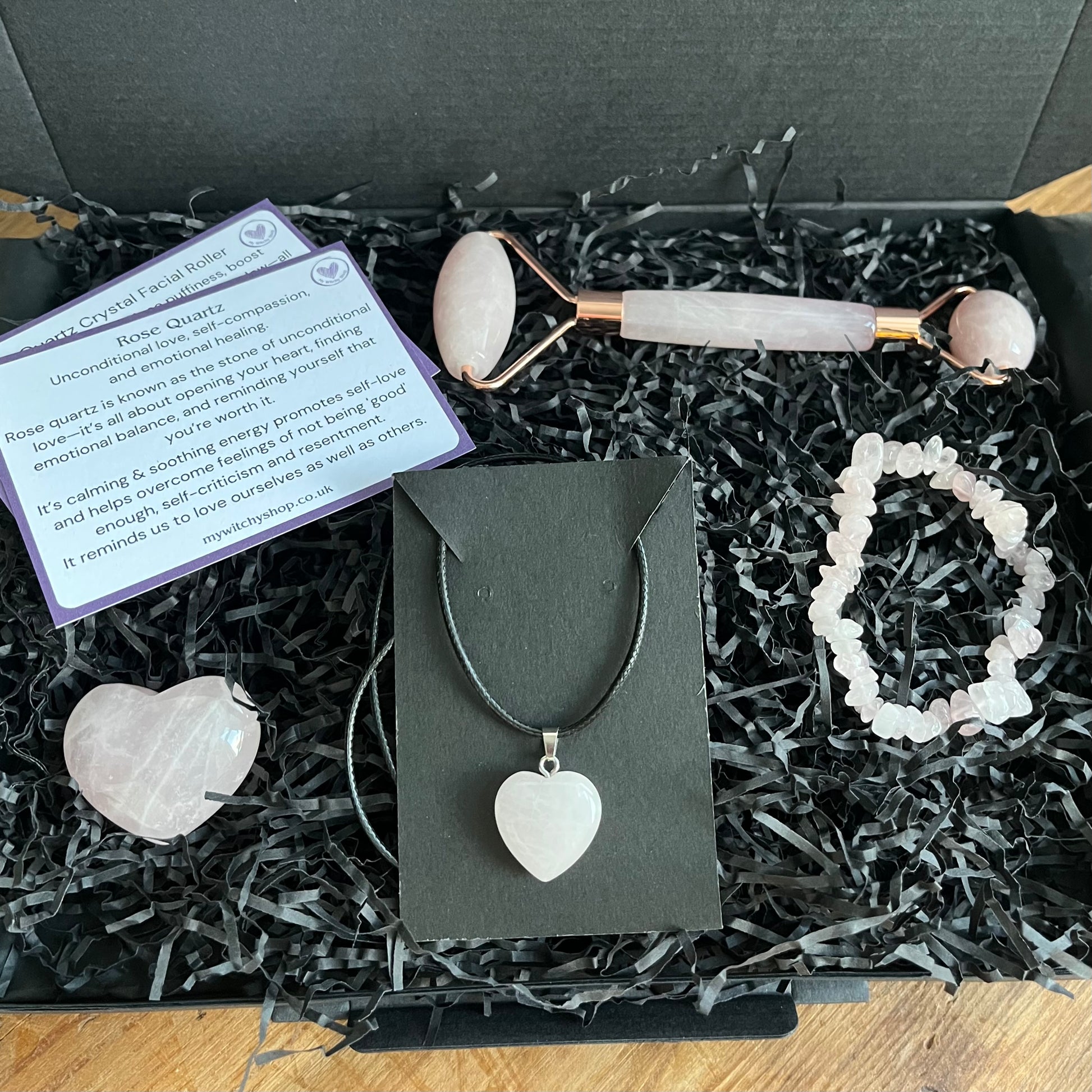 A black Gift box containing a selection of rose quartz items, a rose quartz face roller, rose quartz chip bracelet, a rose quartz heart , and a rose quartz pendent heart necklace. Plus information cards with the properties of rose quartz and benefits of a face roller. 