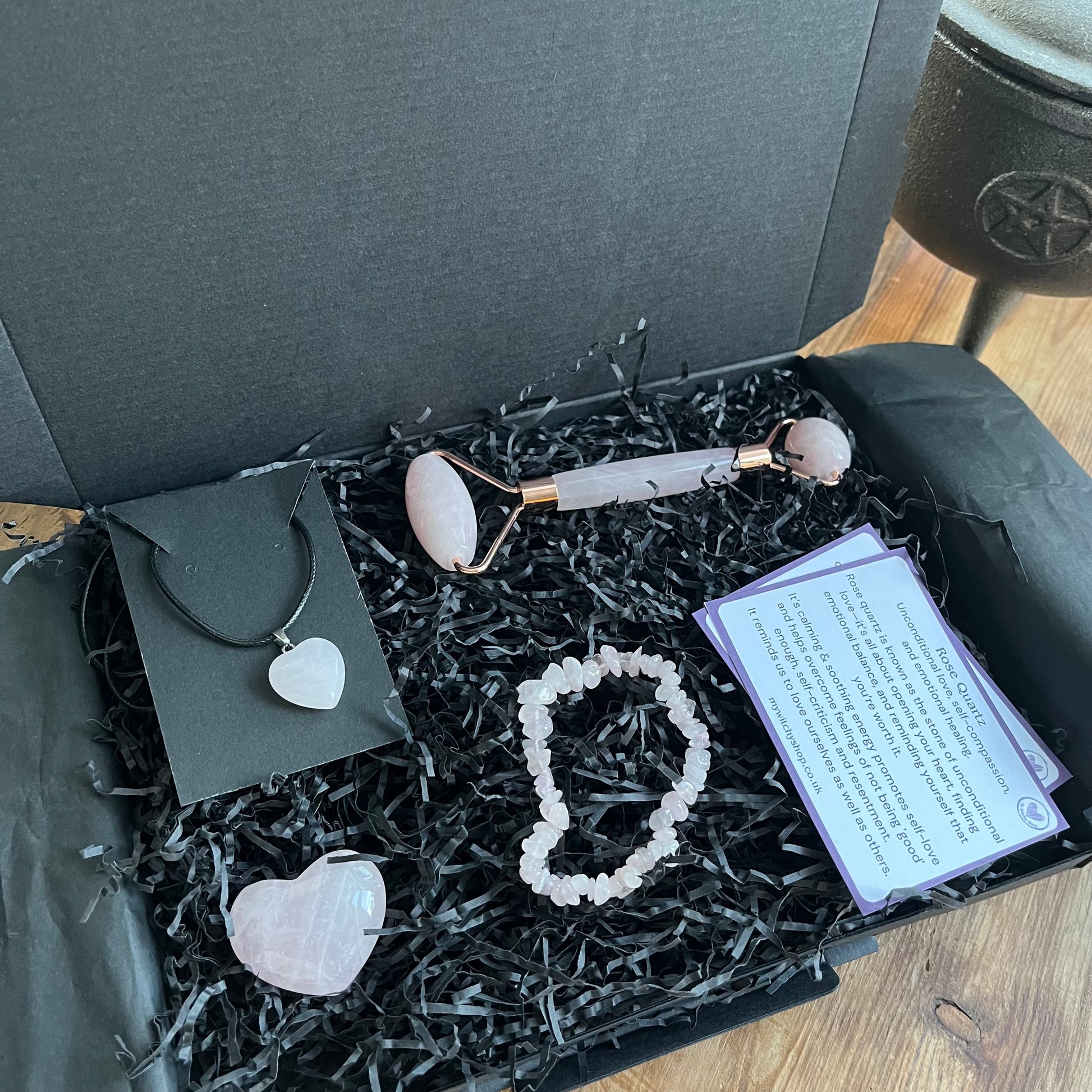 A black gift bow with the contents of the Rose Quartz selfcare gift box. A rose quartz face roller, rose quartz chip bracelet, a rose quartz heart , and a rose quartz pendent heart necklace. Plus information cards with the properties of rose quartz and benefits of a face roller. 