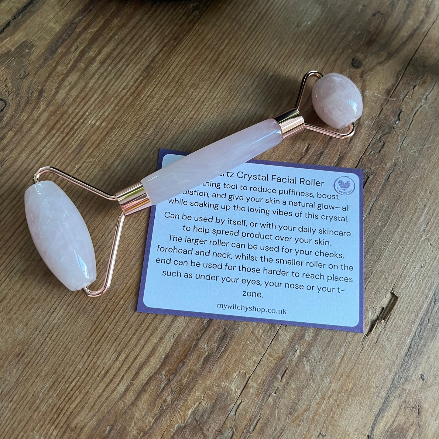 One of the items from the Rose Quartz selfcare gift box sat on a wooden table.  A rose quartz face roller, Plus information card with the properties of rose quartz and benefits of a face roller. 