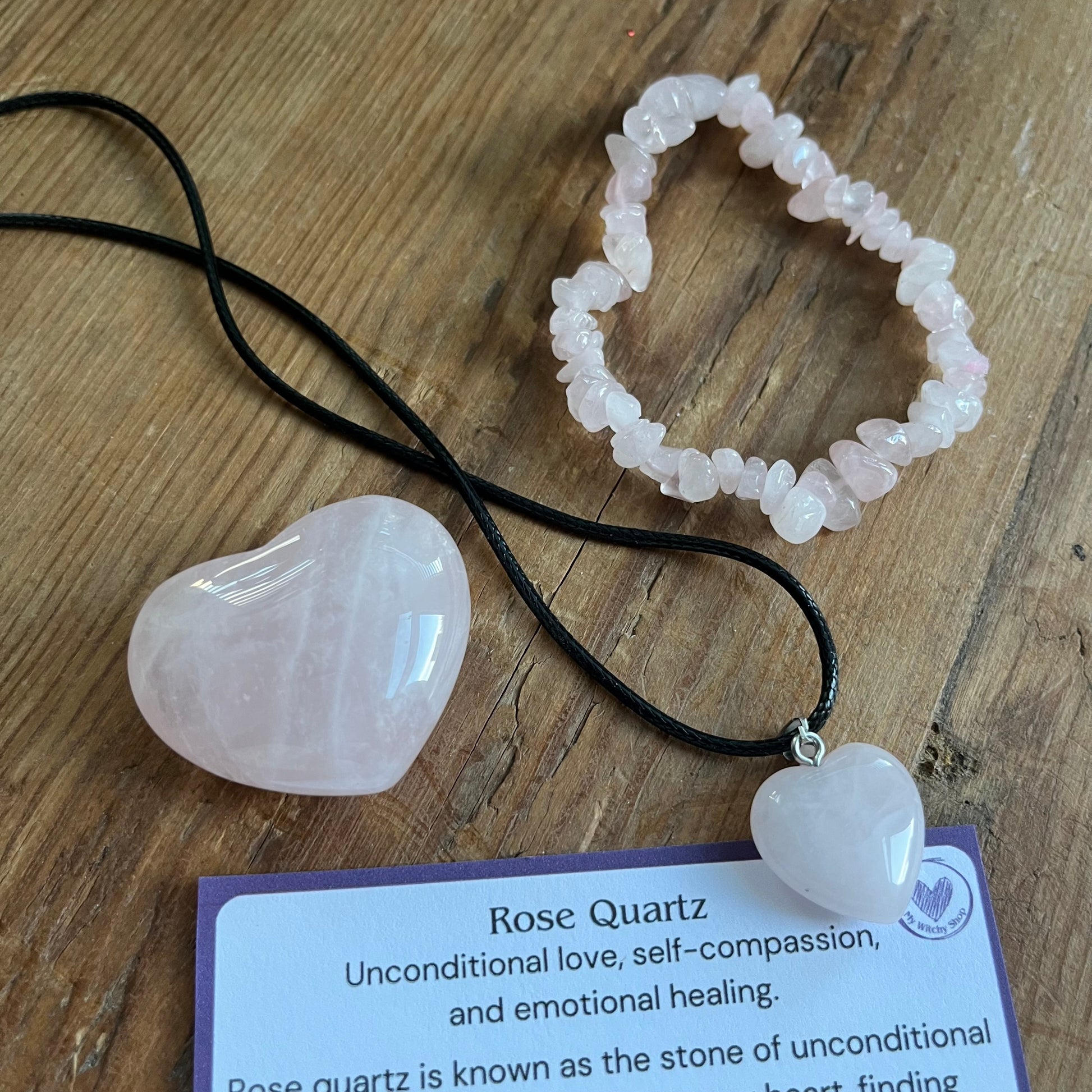 Some of the contents of the Rose Quartz selfcare gift box sat on a wooden table  a rose quartz chip bracelet, a rose quartz heart , and a rose quartz pendent heart necklace. Plus information cards with the properties of rose quartz