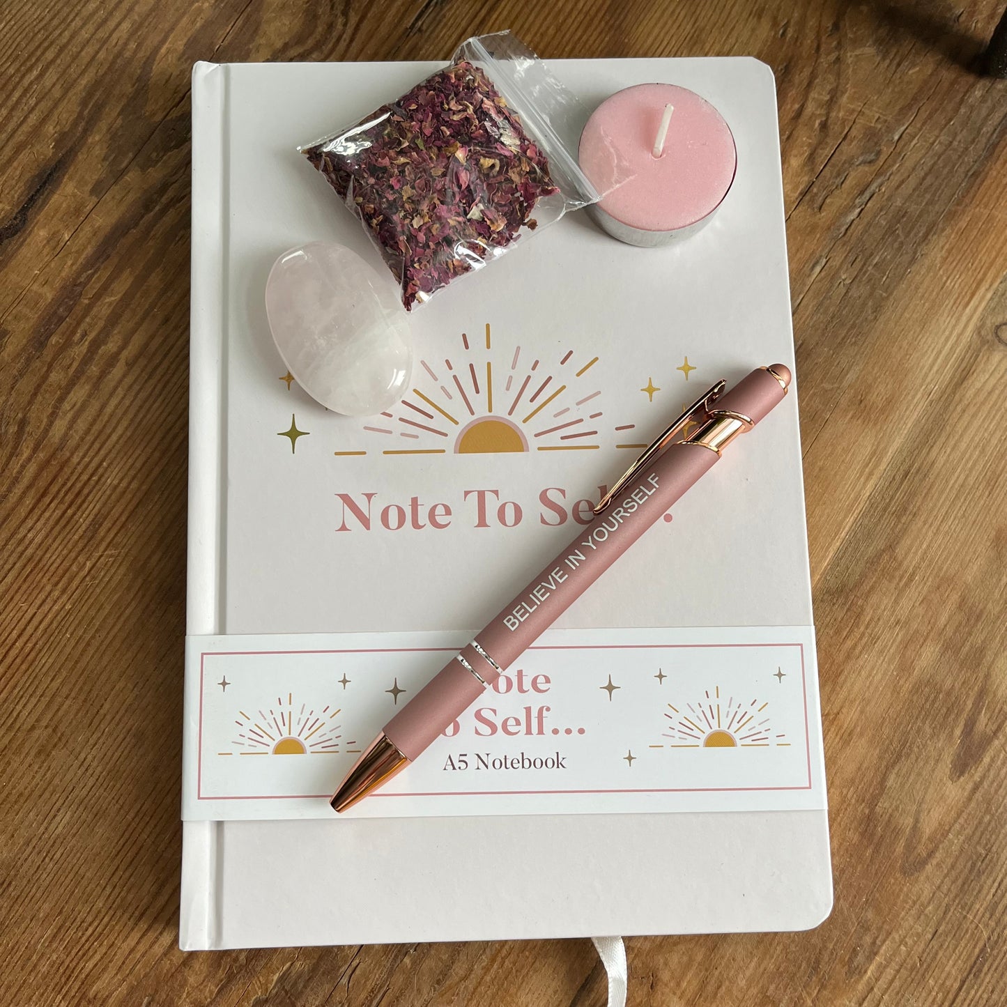 contents of Note to self - ritual for self -love gift box. Pale pink & gold A5 notebook with the words note to self, a rose gold coloured pen with a motivational message written on the side, a rose quartz palm stone, a pink tealight candle and a packet of red rose petals. 