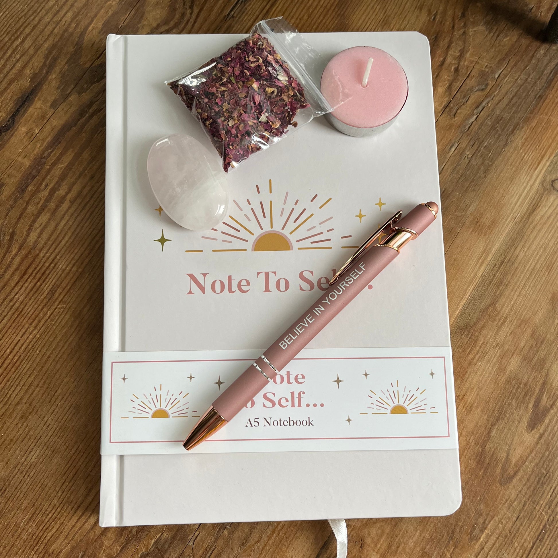 contents of Note to self - ritual for self -love gift box. Pale pink & gold A5 notebook with the words note to self, a rose gold coloured pen with a motivational message written on the side, a rose quartz palm stone, a pink tealight candle and a packet of red rose petals. 