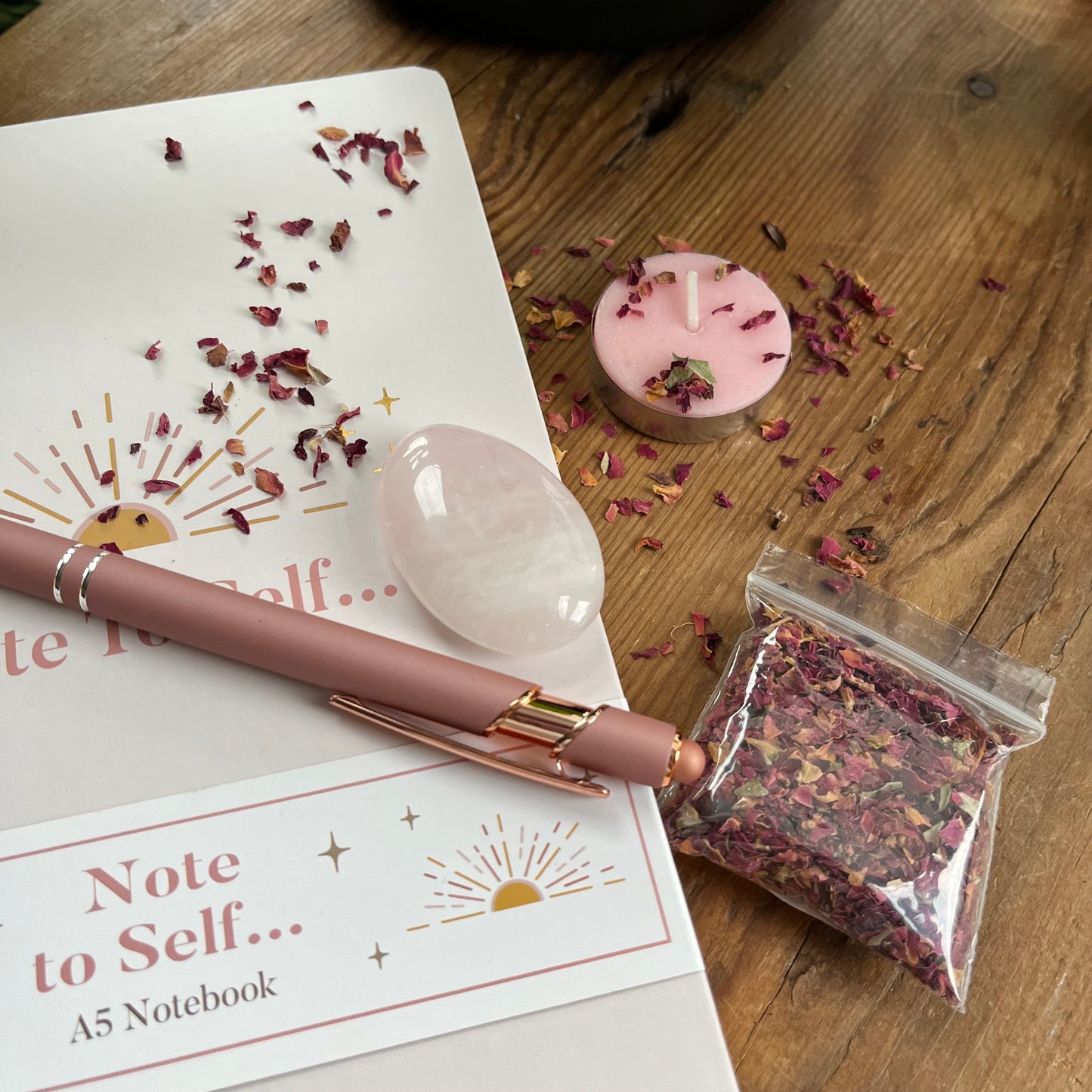 contents of Note to self - ritual for self -love gift box. Pale pink & gold A5 notebook with the words note to self, a rose gold coloured pen with a motivational message written on the side, a rose quartz palm stone, a pink tealight candle and a packet of red rose petals. Plus a parchment style A4 sheet with the ritual guide. The items are sat on a wooden table, rose petals are scattered on and around the candle and notebook as if preparing for a ritual. 