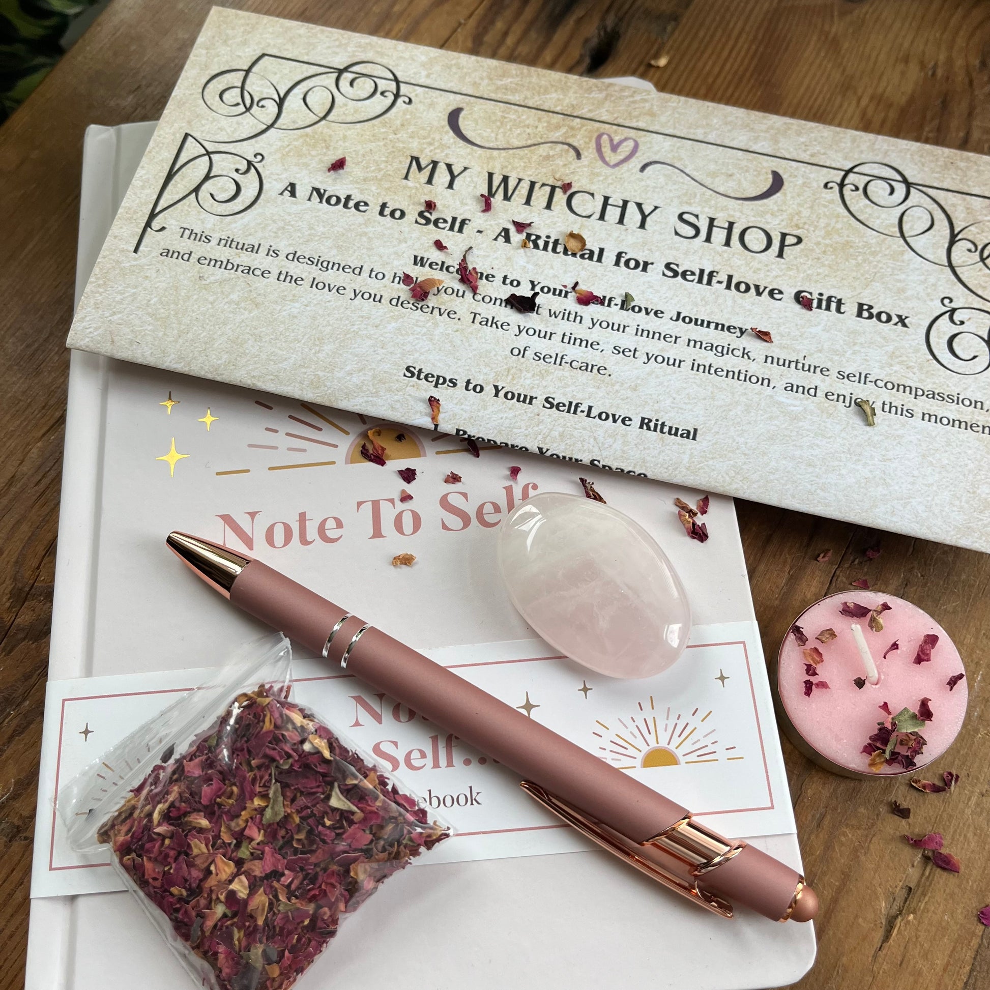 contents of Note to self - ritual for self -love gift box. Pale pink & gold A5 notebook with the words note to self, a rose gold coloured pen with a motivational message written on the side, a rose quartz palm stone, a pink tealight candle and a packet of red rose petals. Plus a parchment style A4 sheet with the ritual guide.  All items are positioned on a wooden table with rose petals scattered over them. 