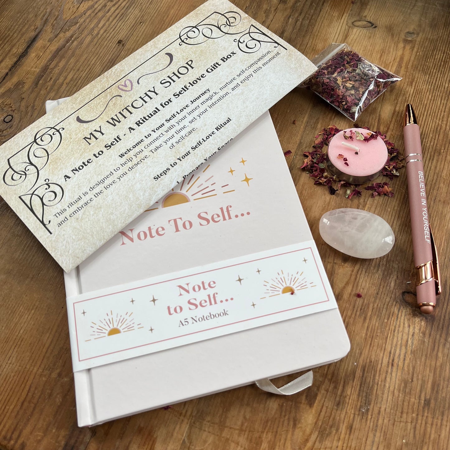 contents of Note to self - ritual for self -love gift box. Pale pink & gold A5 notebook with the words note to self, a rose gold coloured pen with a motivational message written on the side, a rose quartz palm stone, a pink tealight candle and a packet of red rose petals. Plus a parchment style A4 sheet with the ritual guide. 