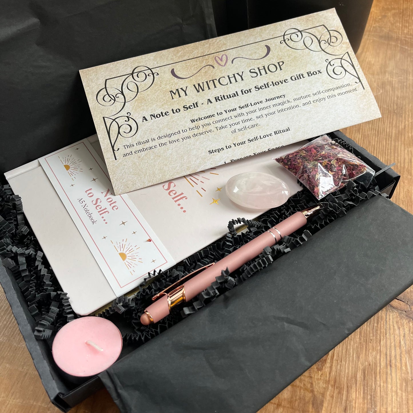 A black letterbox size gift box containing the contents of Note to self - ritual for self -love gift box. Pale pink & gold A5 notebook with the words note to self, a rose gold coloured pen with a motivational message written on the side, a rose quartz palm stone, a pink tealight candle and a packet of red rose petals. Plus a parchment style A4 sheet with the ritual guide. 