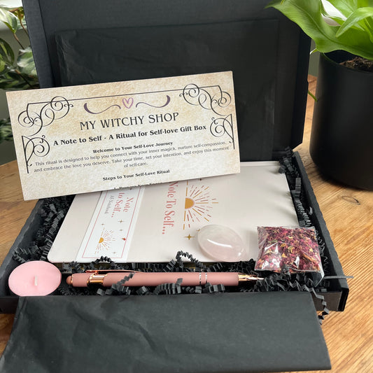 A black letterbox size gift box containing the contents of Note to self - ritual for self -love gift box. Pale pink & gold A5 notebook with the words note to self, a rose gold coloured pen with a motivational message written on the side, a rose quartz palm stone, a pink tealight candle and a packet of red rose petals. Plus a parchment style A4 sheet with the ritual guide. 