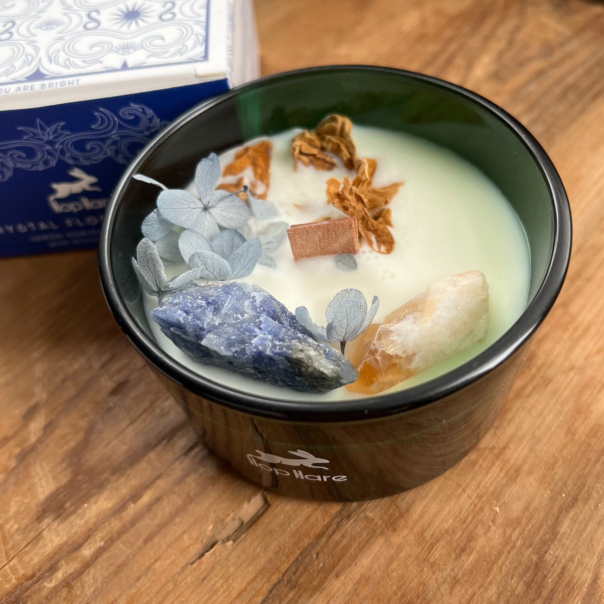 Hop Hare Sun Candle with wooden wick, blue flowers and arge citrine and sodalite crystals. Candle itself is white and set in a dark green glass jar. 