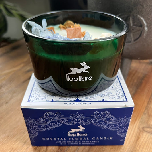 Hop Hare Sun Candle with wooden wick, blue flowers and large citrine and sodalite crystals. Candle itself is white and set in a dark green glass jar. The Candle is shown atop of it's blue and white decorative cardboard presentation box. 