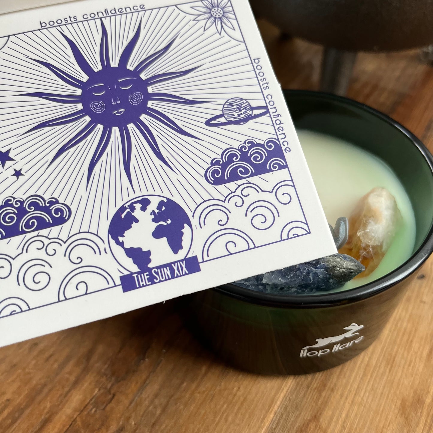 Hop Hare Sun Candle with wooden wick, blue flowers and large citrine and sodalite crystals. Candle itself is white and set in a dark green glass jar. The Candle is shown nest to its accompanying sun tarot card.