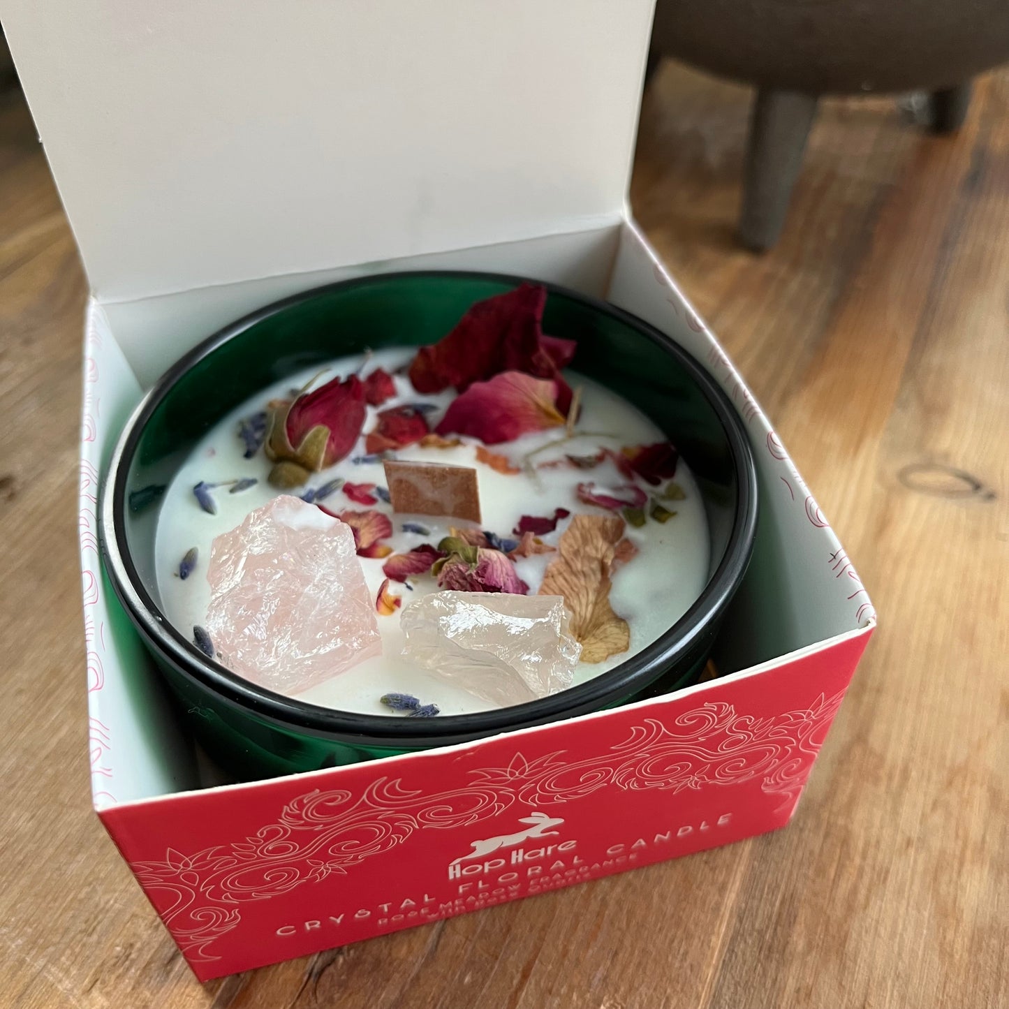 Top of the hop hare Lovers candle shows a wooden wick, large rose quartz crystals, red rose bud and rose petals. The candle itself is white and set in a dark green glass Jar. The candle is shown in its decorated cardboard presentation box 