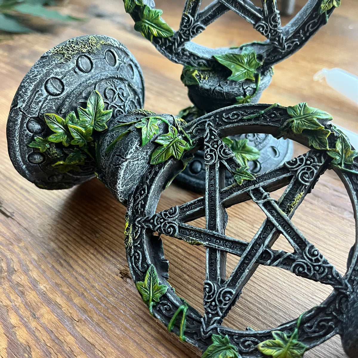 A close-up of a decorative pentagram figurine with a weathered dark gray and black finish, detailed with faux green ivy leaves, resting on a wooden surface.