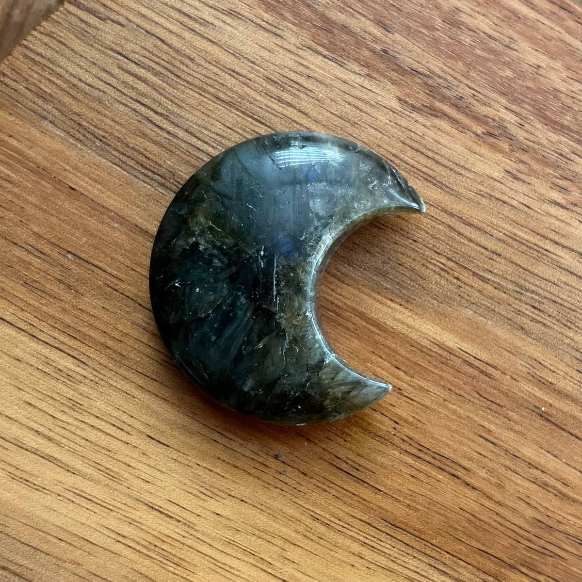 A polished crescent moon-shaped crystal pendant made of labradorite, resting on a wooden surface.