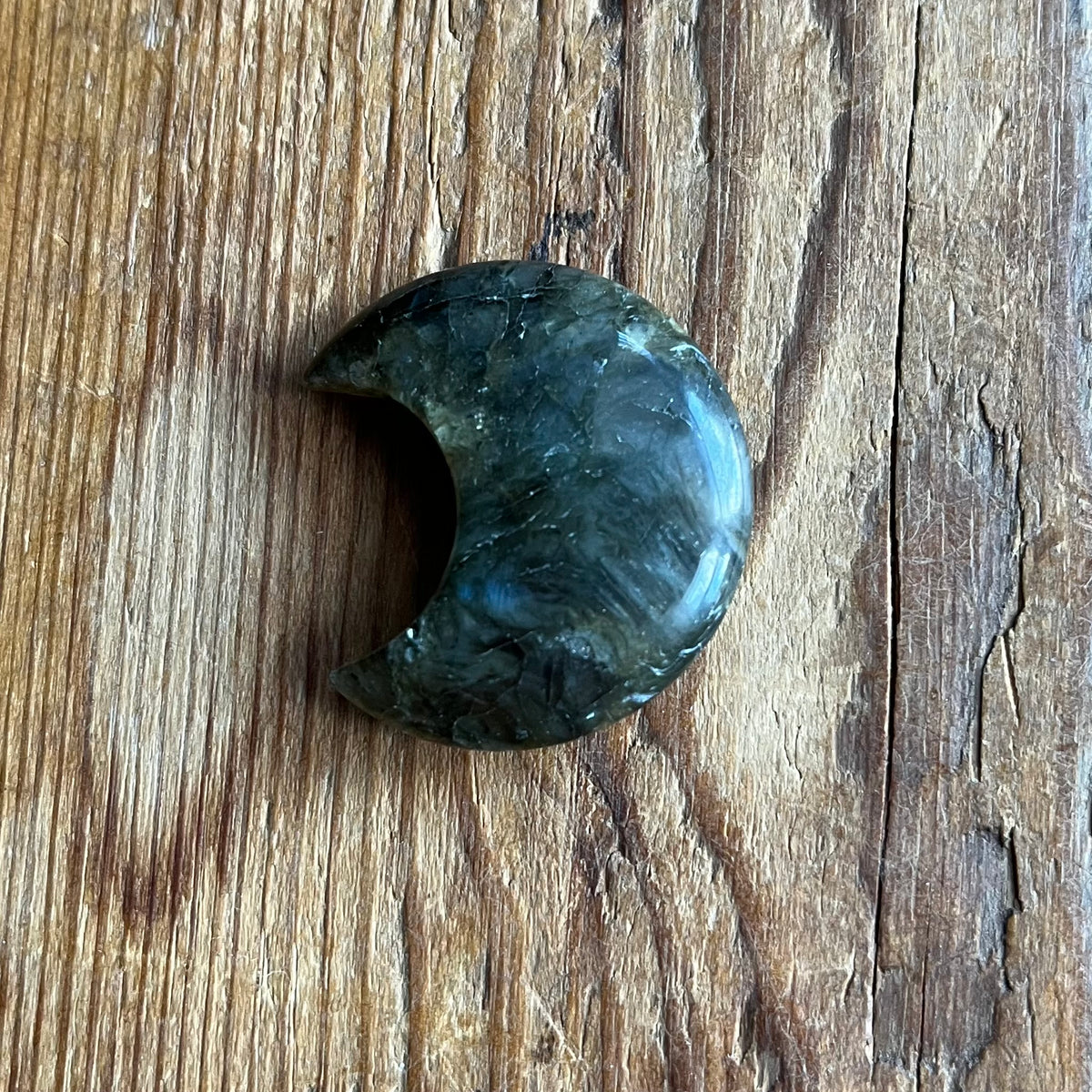 A polished crescent moon-shaped labradorite stone charm resting on a rustic wooden surface.