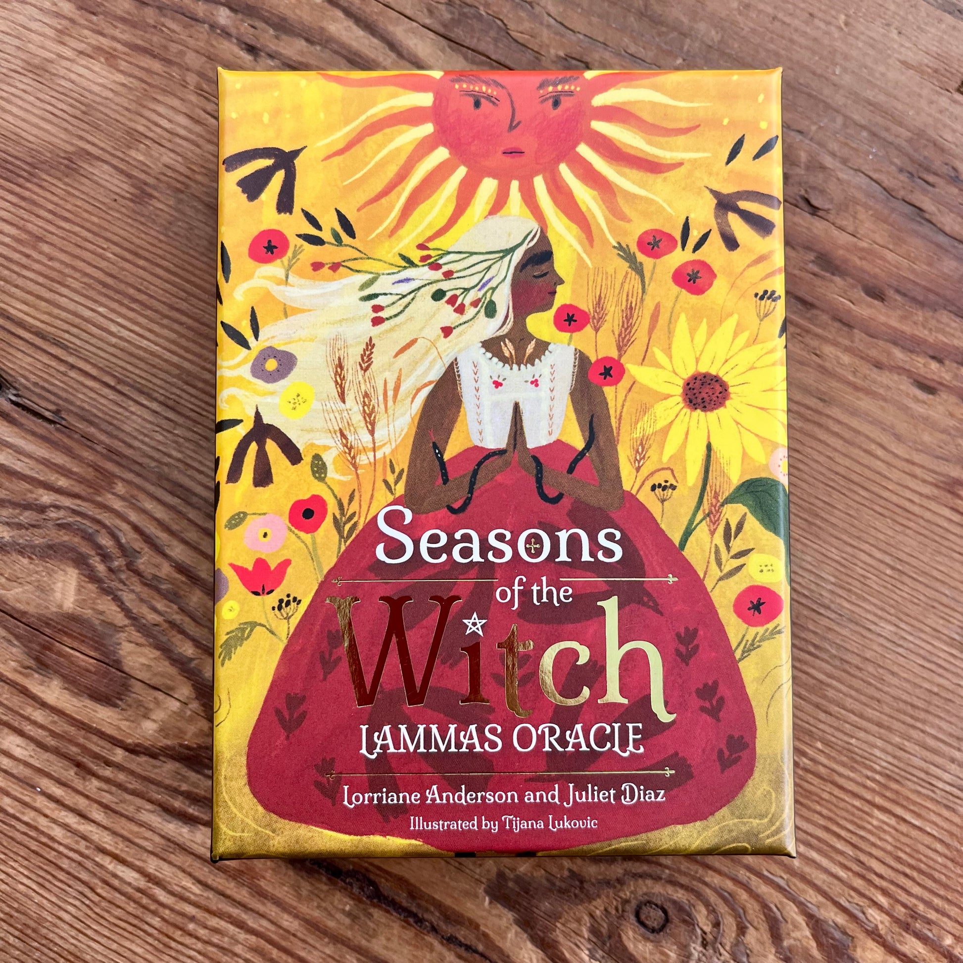 The front cover of the 'Seasons of the Witch Lammas Oracle' card deck by Lorriane Anderson and Juliet Diaz, featuring a stylized illustration of a witch in a red dress under a sun, placed on a wooden surface.
