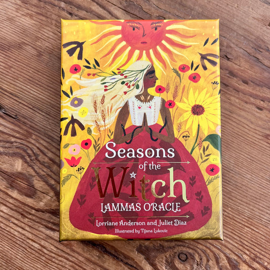 The front cover of the 'Seasons of the Witch Lammas Oracle' card deck by Lorriane Anderson and Juliet Diaz, featuring a stylized illustration of a witch in a red dress under a sun, placed on a wooden surface.