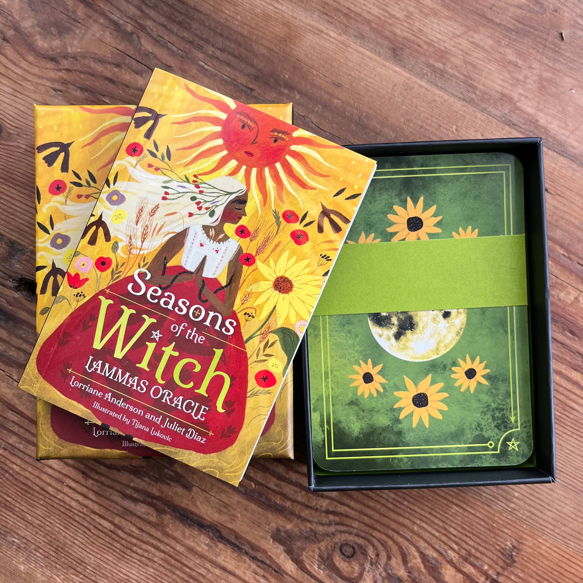 Witch's Oracle card deck and box set on a wooden tabletop. The front of the box shows the title 'Seasons of the Witch' with illustrated suns, flowers, and a figure, while the open black box reveals green-backed cards with sun and moon artwork and a green paper band around them.