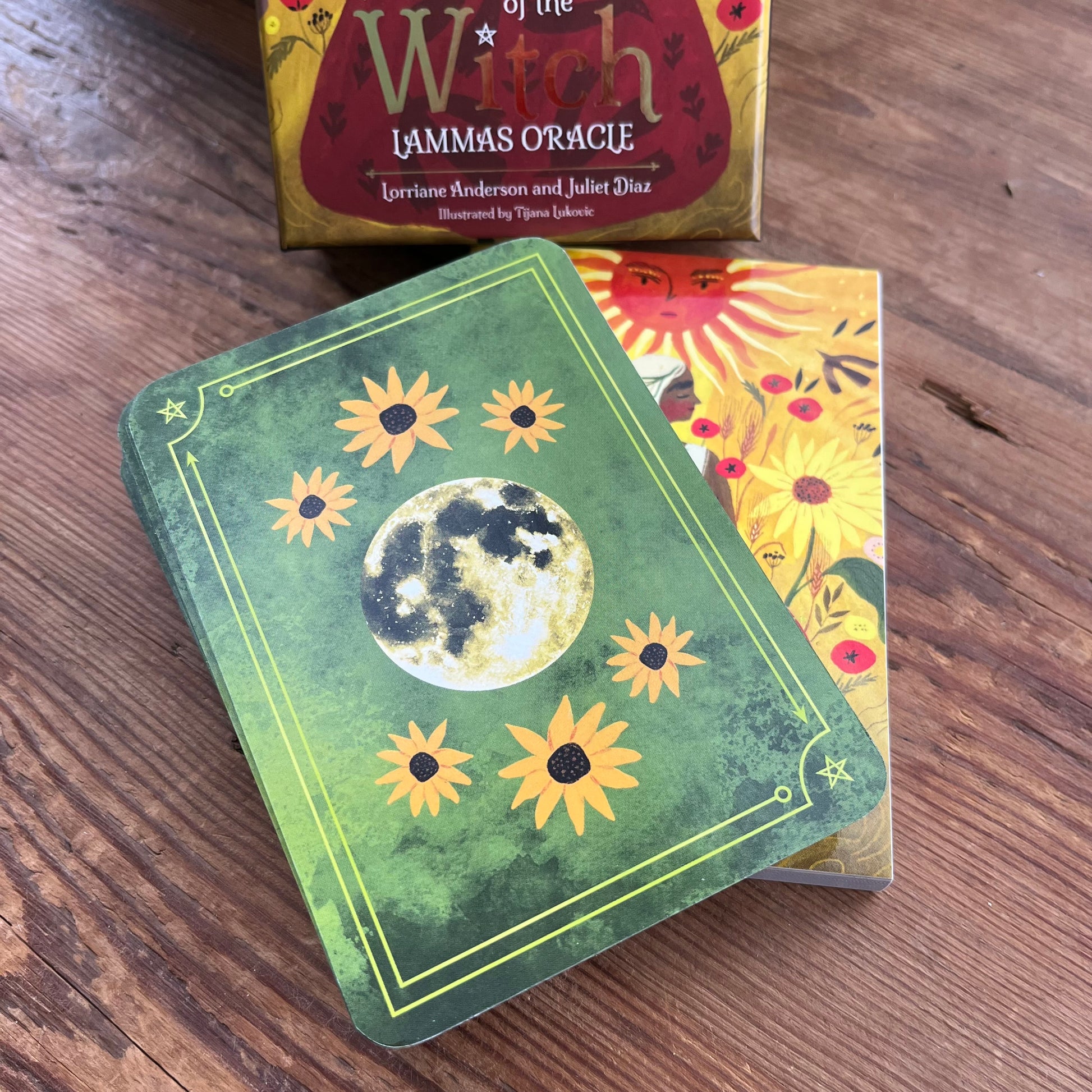 A boxed card deck titled "Witch Lammas Oracle" sits on a wooden table with two illustrated cards in front. The front card is green with a moon illustration and yellow sun motifs, and the box behind it shows additional colorful artwork and title text.