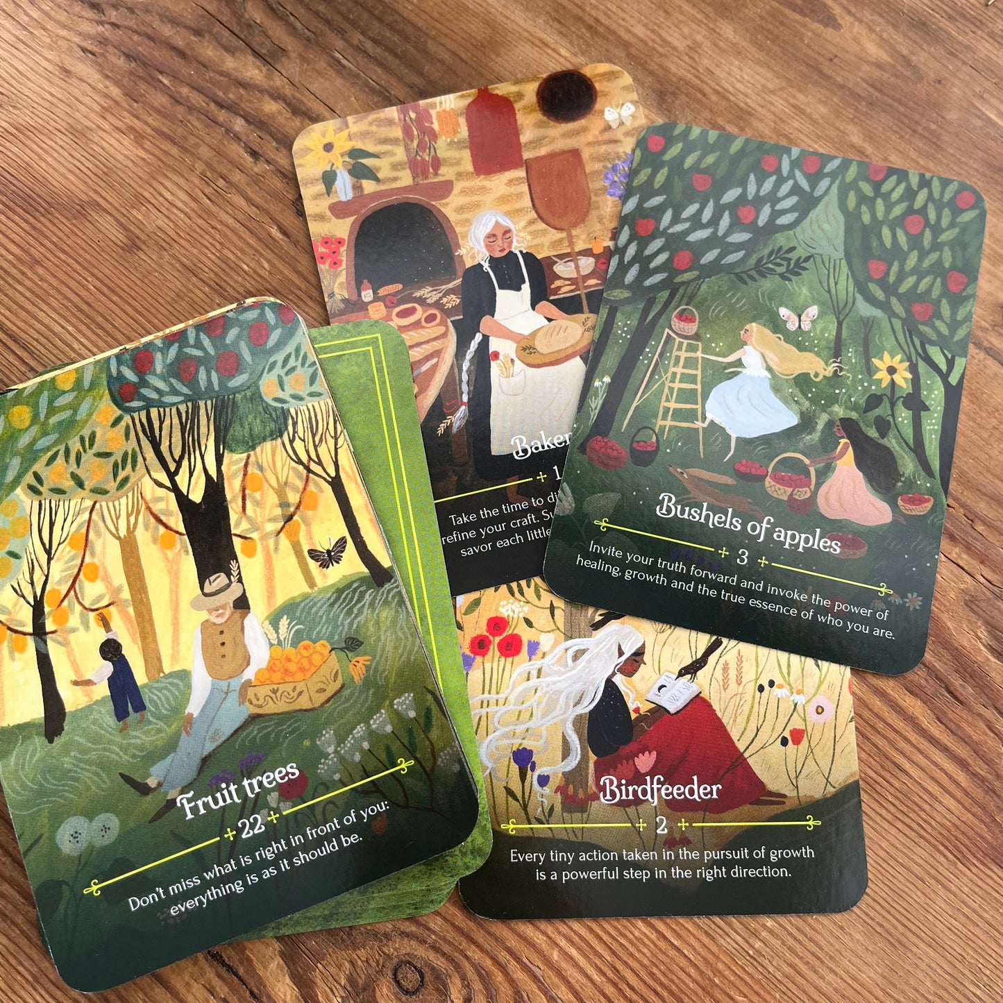 Four illustrated oracle cards fanned out on a wooden tabletop. The cards feature whimsical fantasy and nature scenes with text boxes, including titles like Fruit trees, Bushels of apples, and Birdfeeder.