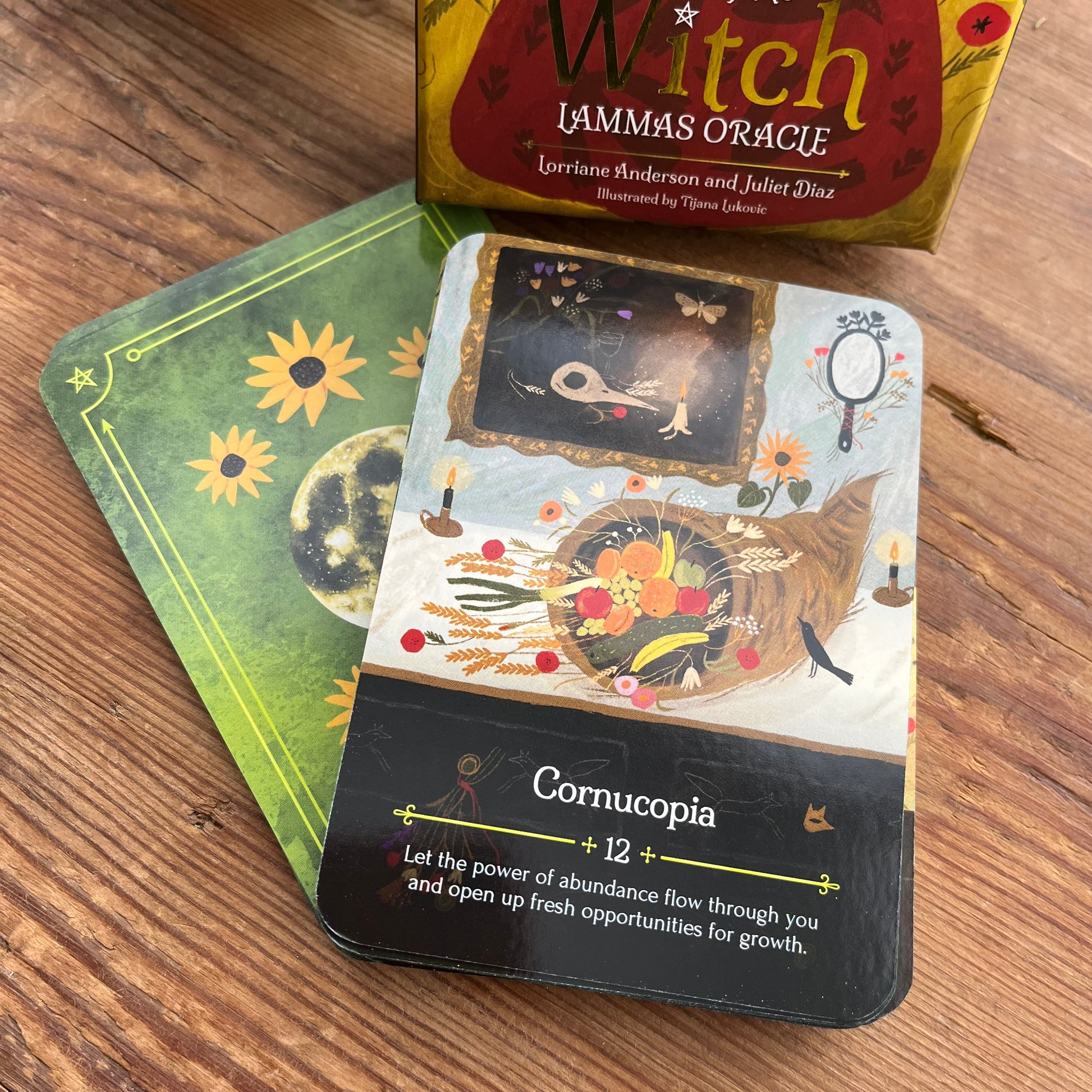 Witch Lammas Oracle card deck on a wooden surface, showing the Cornucopia card, a green card underneath, and the branded box at the top.