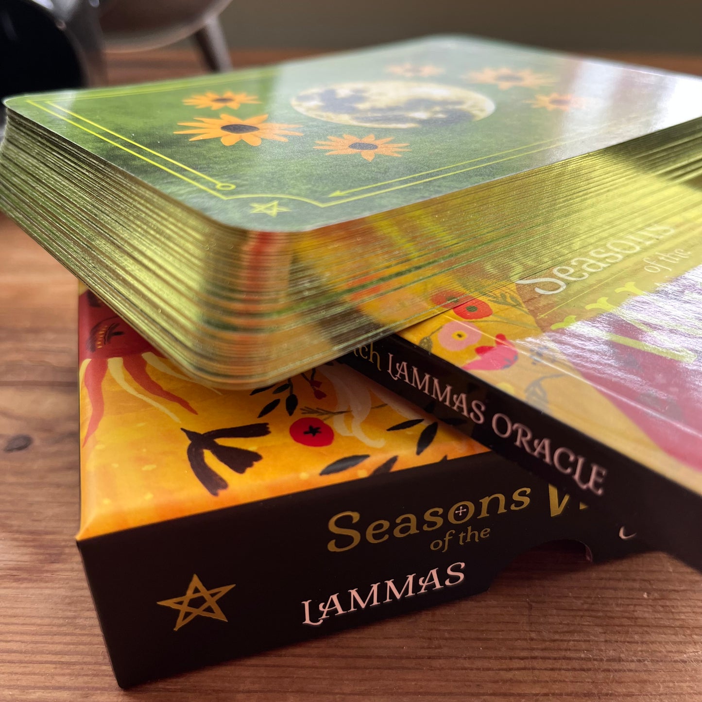 A deck of tarot cards from The Lammas Oracle, with the top card showing a green and gold celestial design, resting on top of its yellow and black illustrated box on a wooden surface.