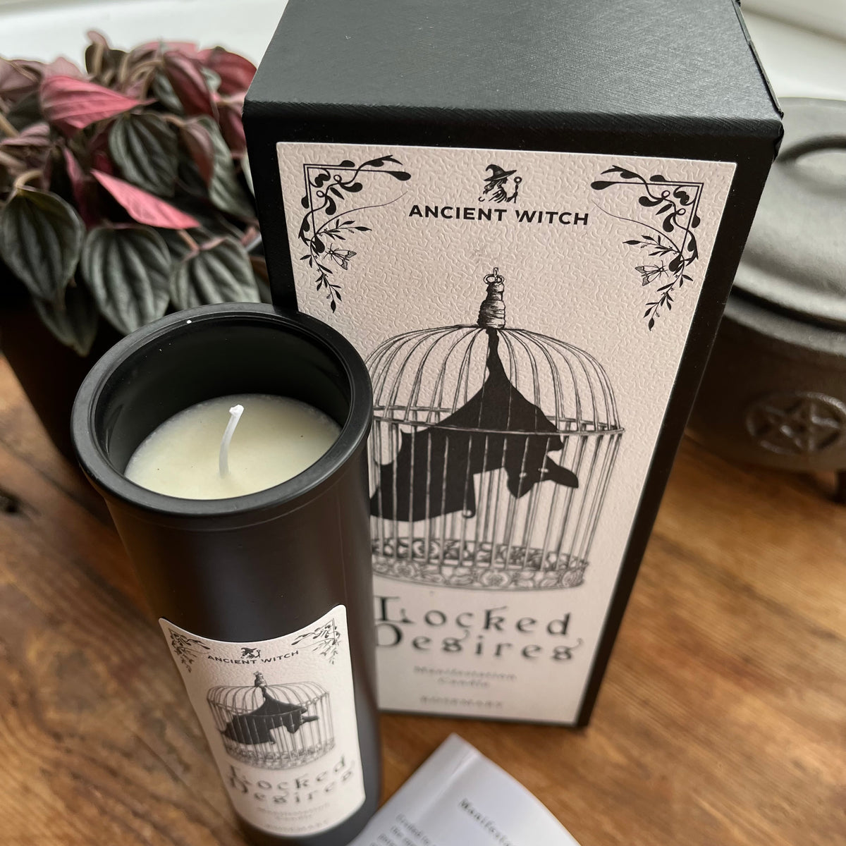 A black candle jar labeled 'Locked Desire' with cream wax sits beside its matching black and cream packaging box on a wooden surface. The box features an illustration of a bird in a cage and the brand name 'Ancient Witch'.