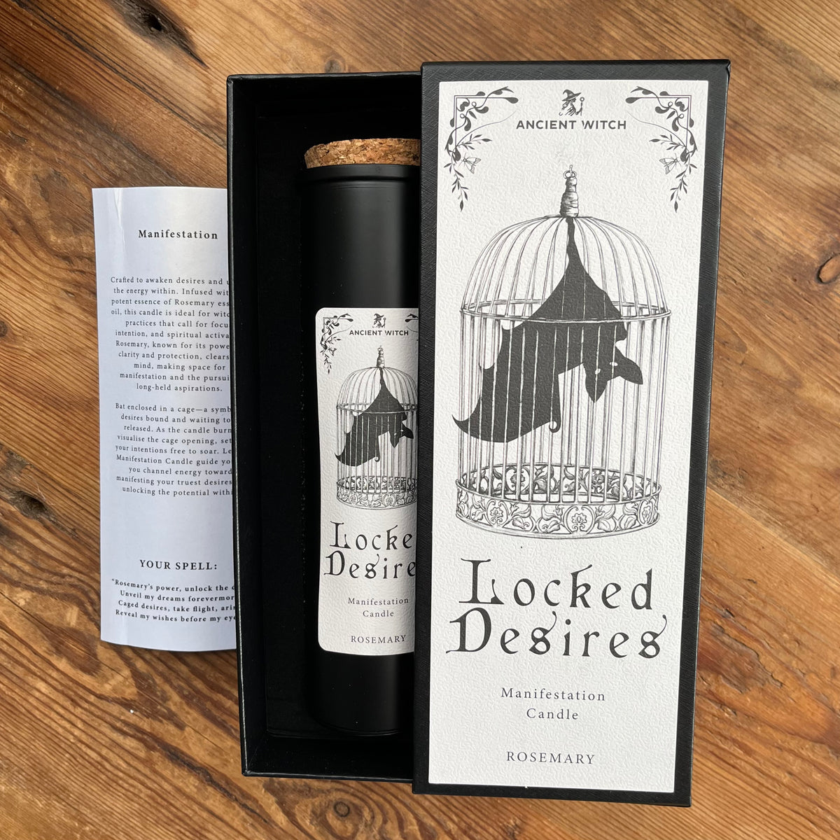 A black cylindrical candle in a black presentation box with a cork stopper, shown beside a printed card and a tall matching label. The label reads 'Ancient Witch Locked Desires Manifestation Candle' and 'Rosemary,' with an illustration of a birdcage and bat.