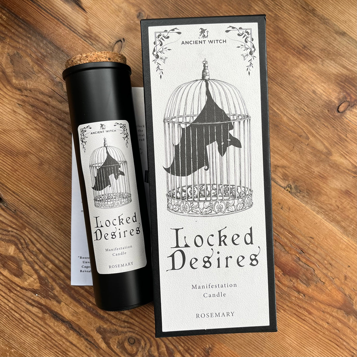 A black cylindrical candle with a cork stopper and a white label reading 'Locked Desires' sits beside its matching rectangular box on a wooden tabletop. The packaging features a black bat inside a birdcage illustration and the brand name 'Ancient Witch'