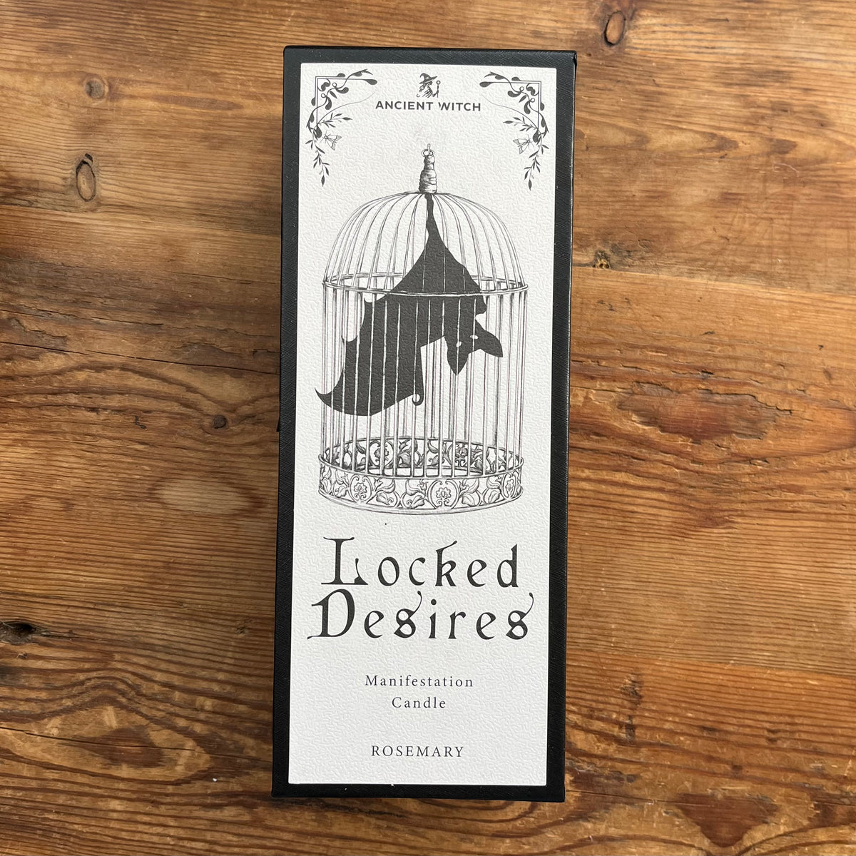 A tall rectangular candle box labeled 'Ancient Witch' and 'Locked Desires Manifestation Candle' with 'Rosemary' at the bottom, placed on a rustic wooden surface. The front shows a black-and-white illustration of a birdcage with a bat inside.