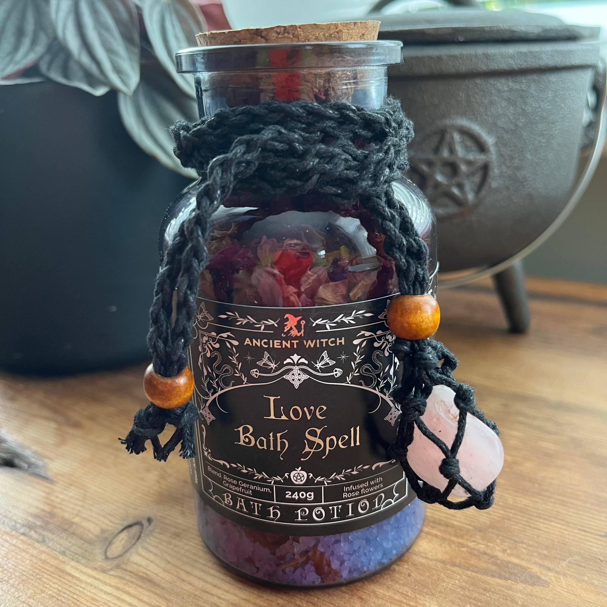 A glass jar labeled 'Ancient Witch Love Bath Spell' filled with purple bath salts and dried flowers, wrapped in black macrame with wooden beads and a pink crystal, set on a wooden surface with a cauldron in the background.