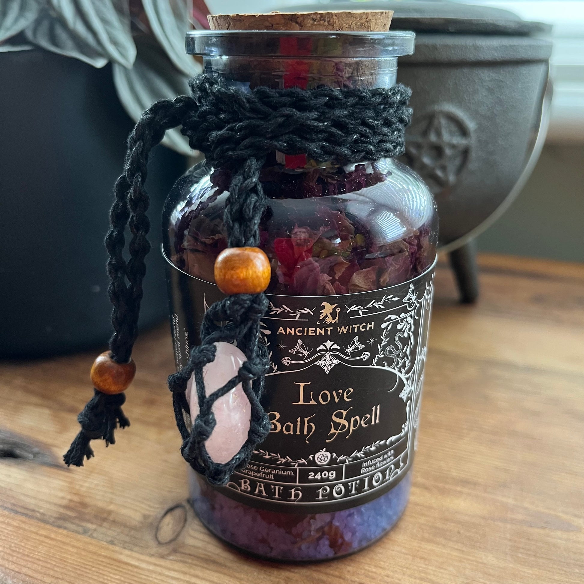 A glass jar labeled 'Love Bath Spell' by Ancient Witch, filled with dried flowers and bath salts, wrapped in a black macrame knot with wooden beads and a pink stone charm, sitting on a wooden surface.