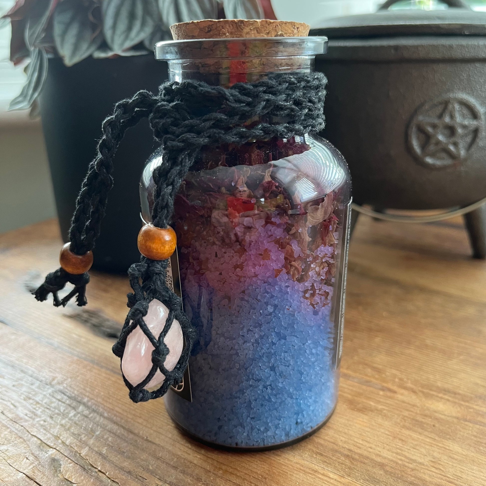 A glass jar candle filled with blue and purple crystals, sealed with a cork and wrapped in a black macrame cord with a pink crystal pendant, set on a wooden table.