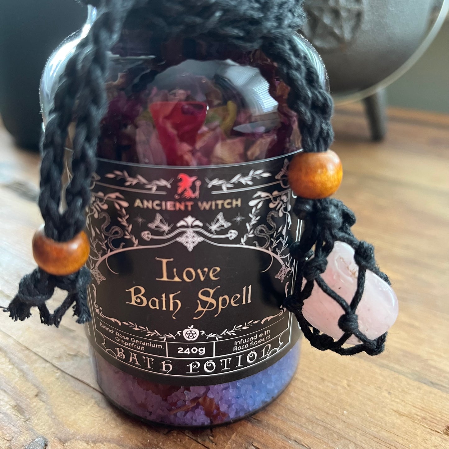 A glass bottle of 'Love Bath Spell' bath potion by Ancient Witch, featuring a black label with gothic lettering, a black hanging cord with wooden beads, and a pink crystal charm, set on a wooden surface.