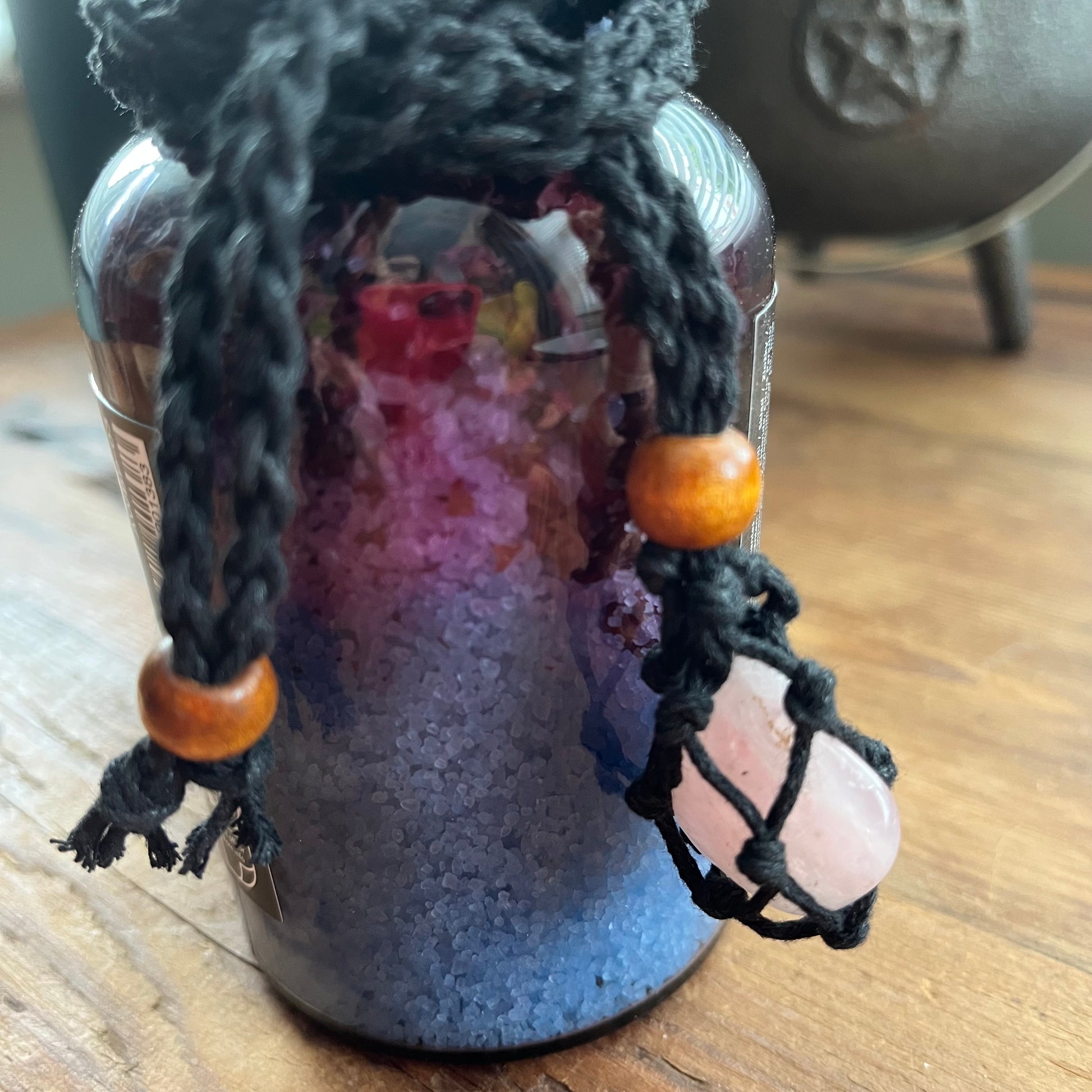 A glass jar filled with purple and blue crystals, decorated with a black macrame hanging charm featuring wooden beads and a pink rose quartz pendant, resting on a wooden surface.