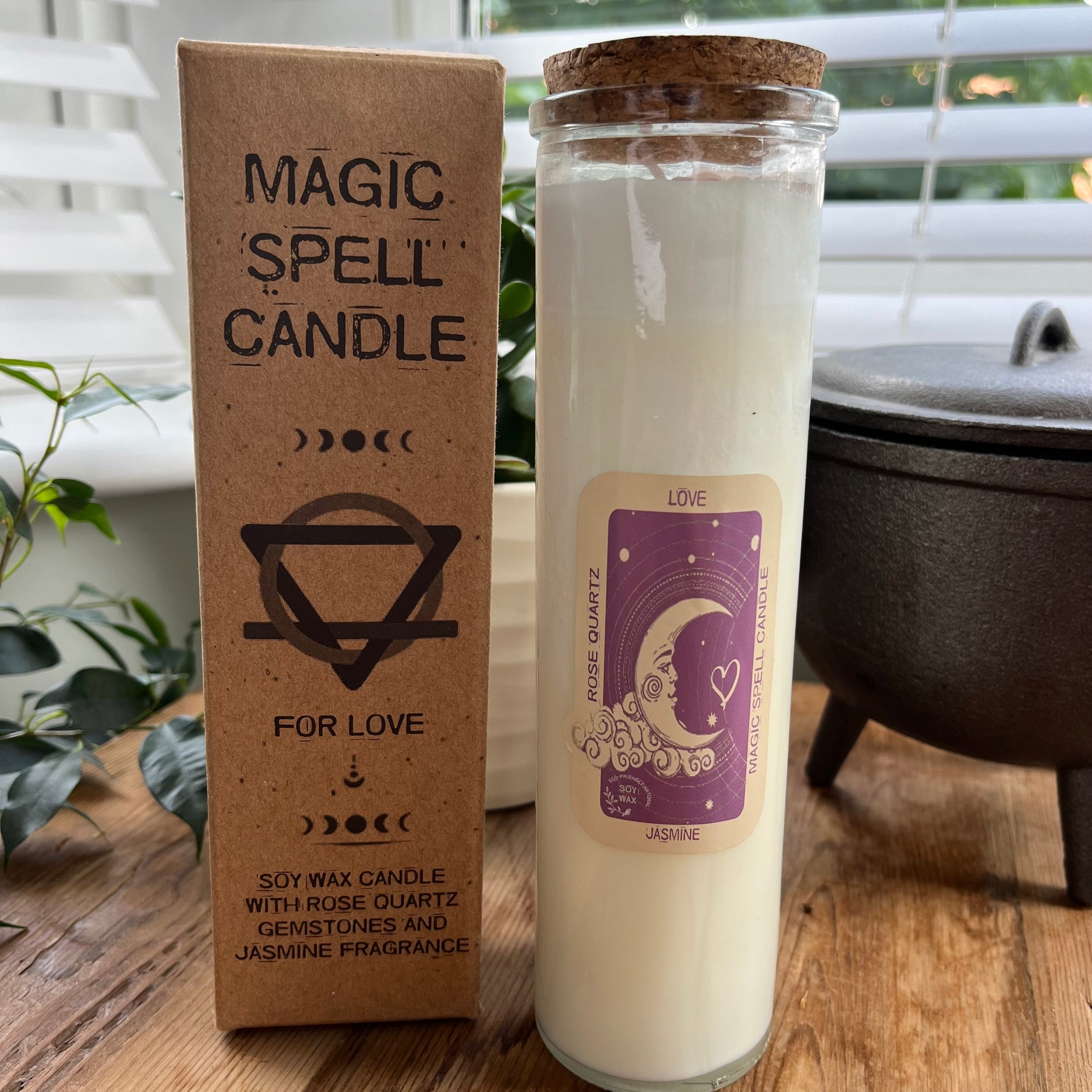 Tall glass spell candle with cork lid. The label on the candle features a crescent moon and love heart and the words Love, rose quartz and Jasmine. The candle itself is white with rose quartz gemstones embedded within it. Alongside the glass Magic spell candle is it's brown cardboard box which has the words magic spell candle for love, soy wax candle with rose quartz gemstones and Jasmine fragrance.