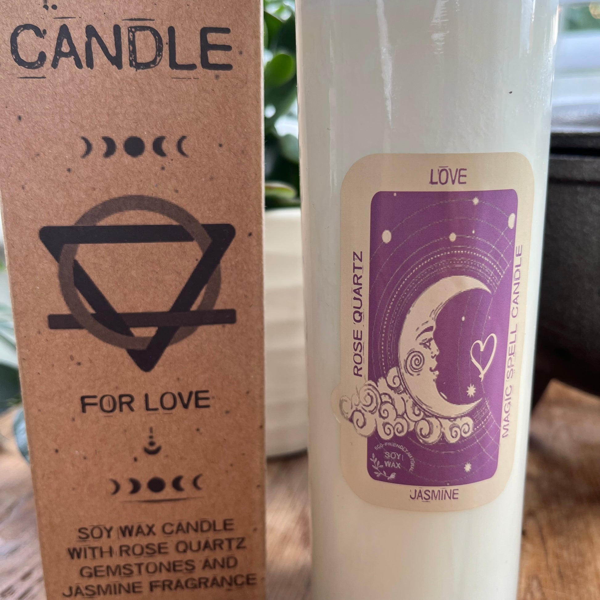 Tall glass spell candle with cork lid. The label on the candle features a crescent moon and love heart and the words Love, rose quartz and Jasmine. The candle itself is white with rose quartz gemstones embedded within it. Alongside the glass Magic spell candle is it's brown cardboard box which has the words magic spell candle for love, soy wax candle with rose quartz gemstones and Jasmine fragrance.