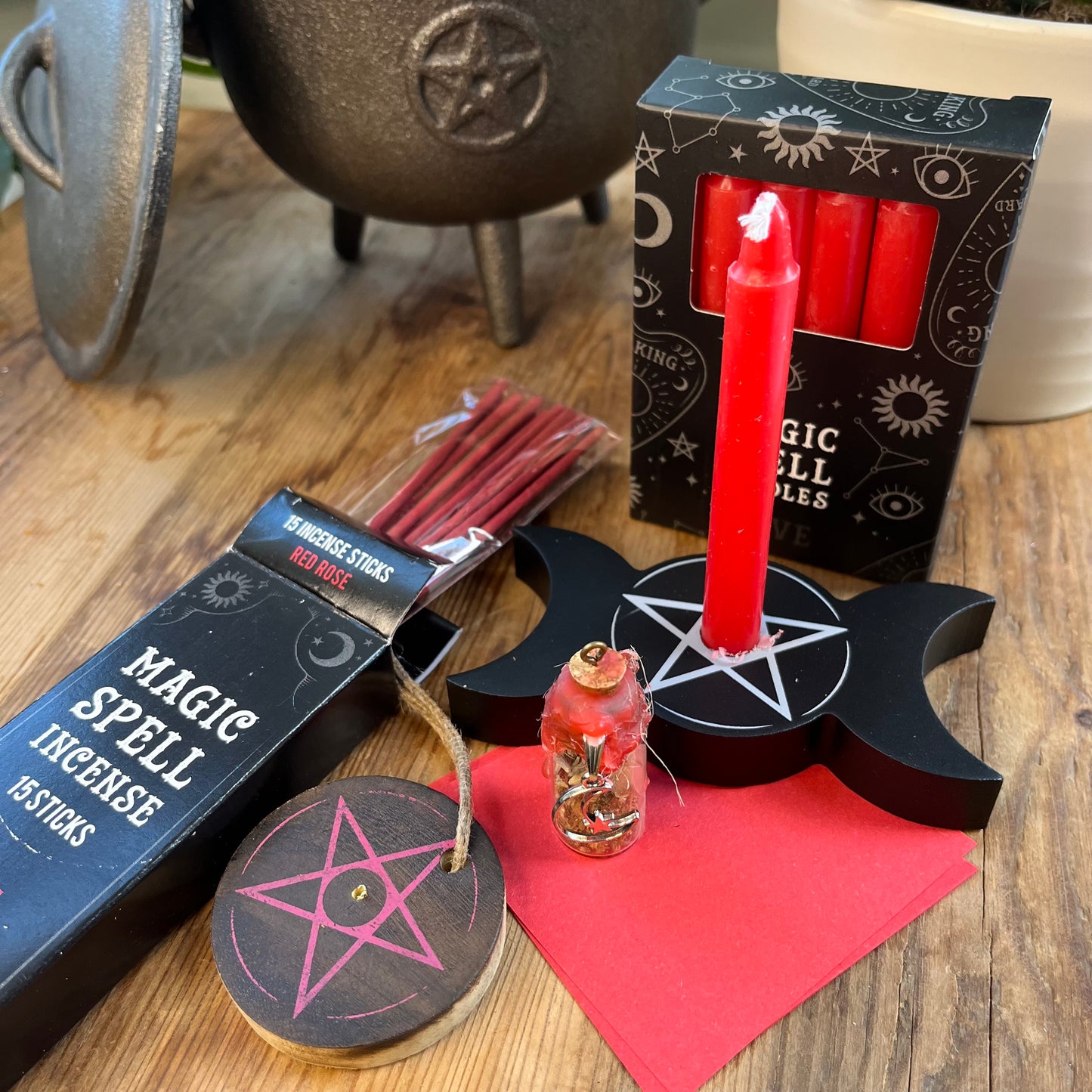 'Love Spell Kit' Contents displayed on a wooden table. Pack of Love Incense sticks, and wooden pentagram incense holder, Pack of red magic spell candles, wooden black candle holder with pentagram design, squares of red paper and a "Love' spell bottle containing herbs and sealed with red wax and a charm.