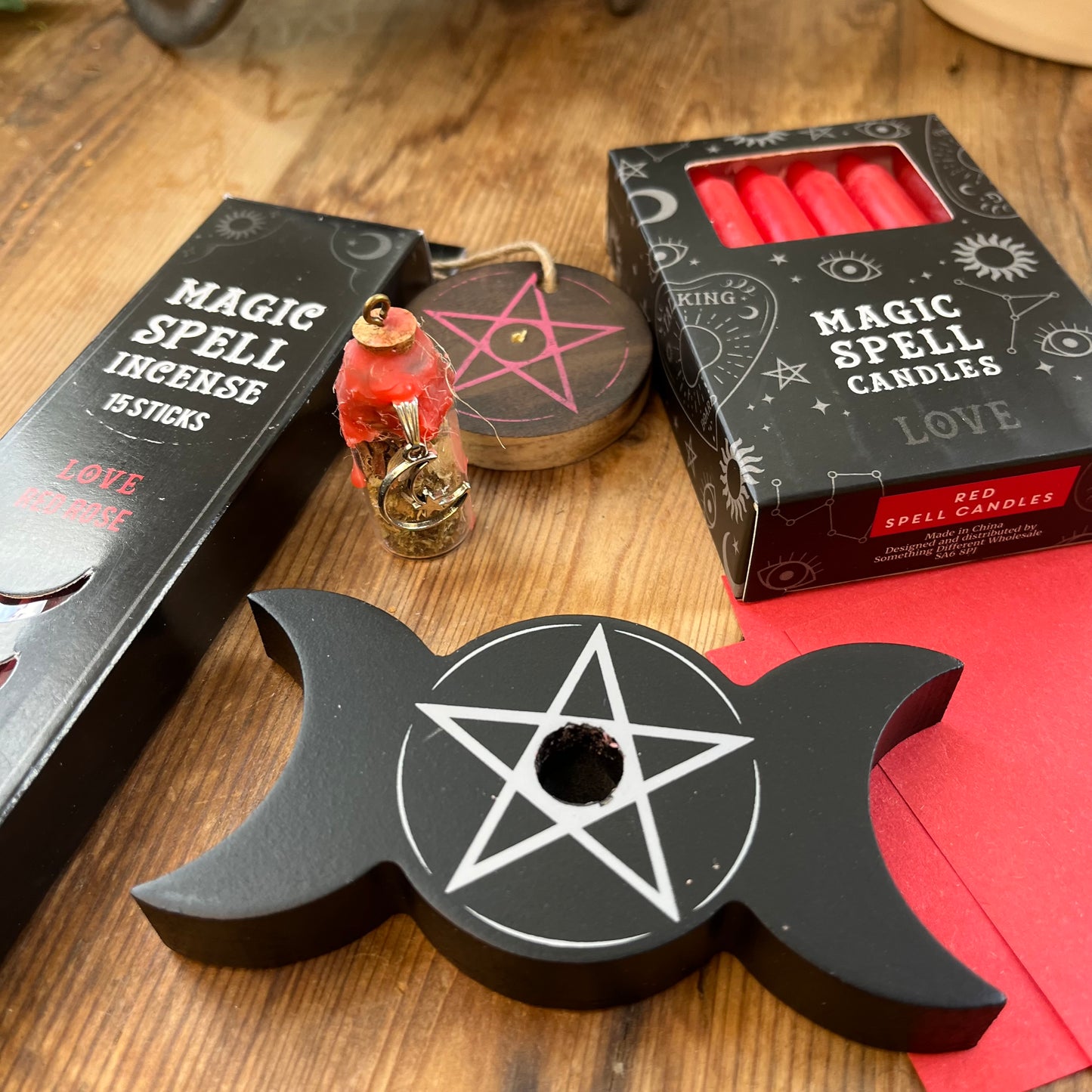 'Love Spell Kit' Contents displayed on a wooden table. Pack of Love Incense sticks, and wooden pentagram incense holder, Pack of red magic spell candles, wooden black candle holder with pentagram design, squares of red paper and a "Love' spell bottle containing herbs and sealed with red wax and a charm.