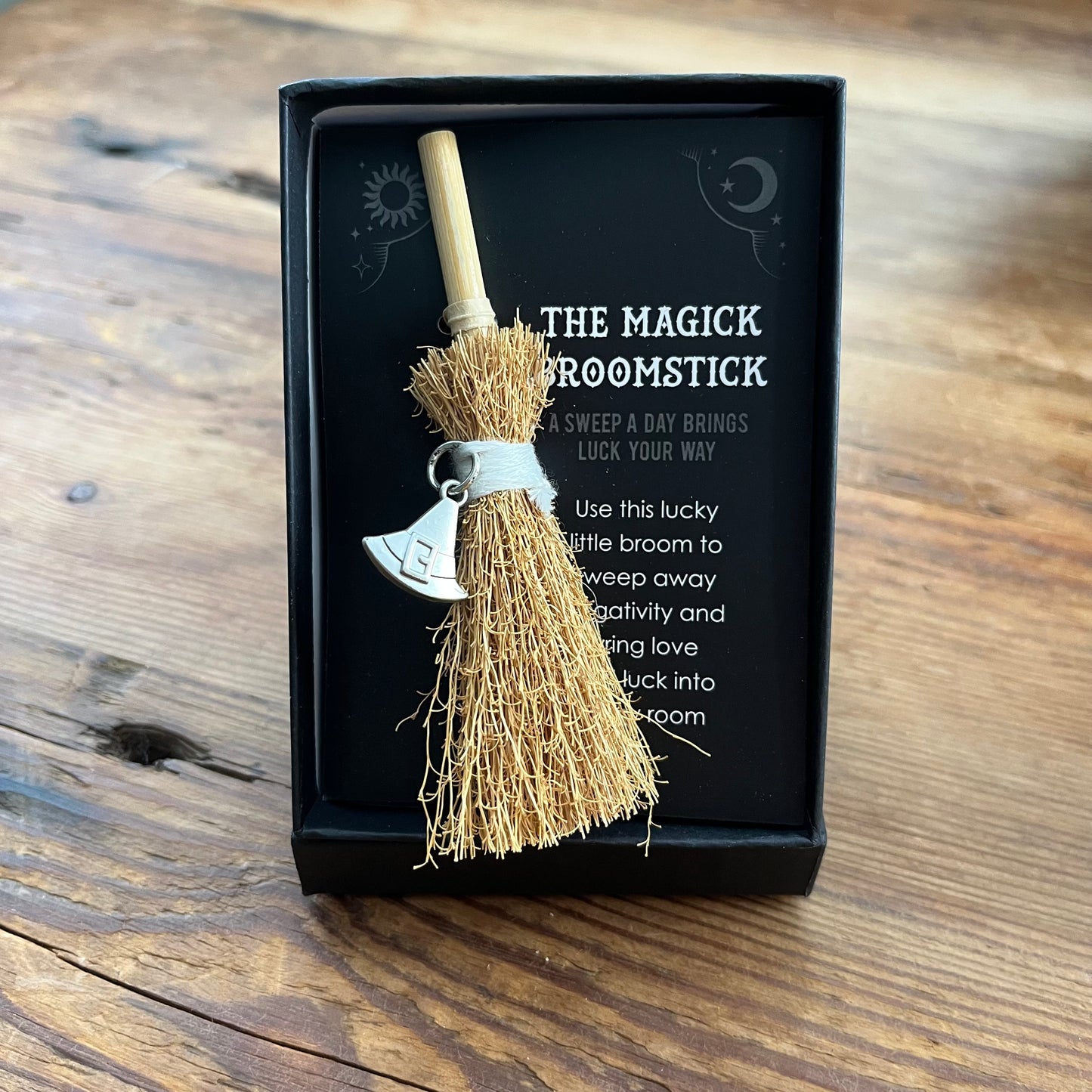 A miniature wooden broomstick charm with a silver witch's hat charm, presented in a black gift box on a rustic wooden table. The box features a printed insert with a sun and moon graphic and the text 'THE MAGICK BROOMSTICK'.
