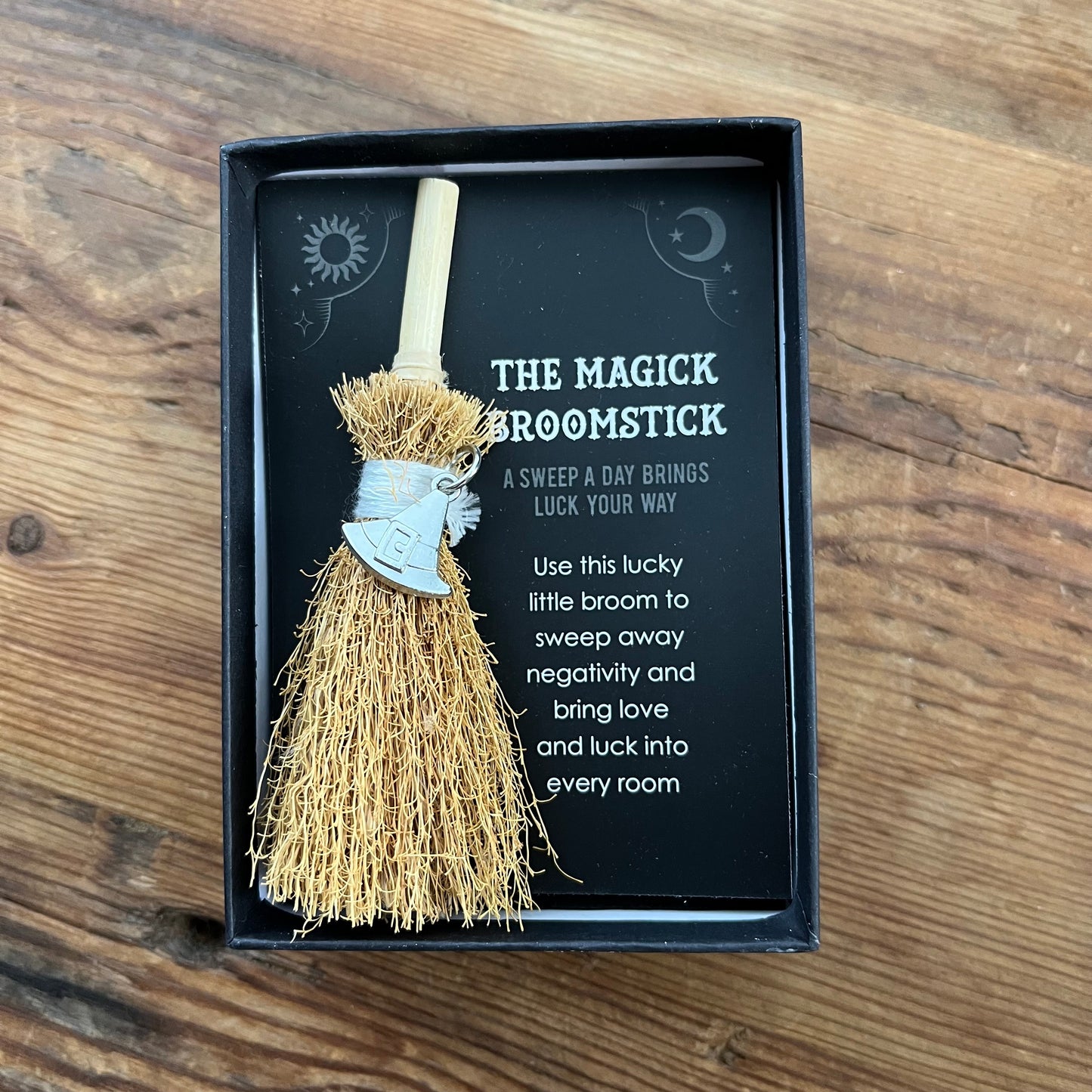 A small decorative broomstick gift in a black presentation box on a wooden surface. The broom has a light wood handle, tan bristles, and a silver metal witch's hat charm