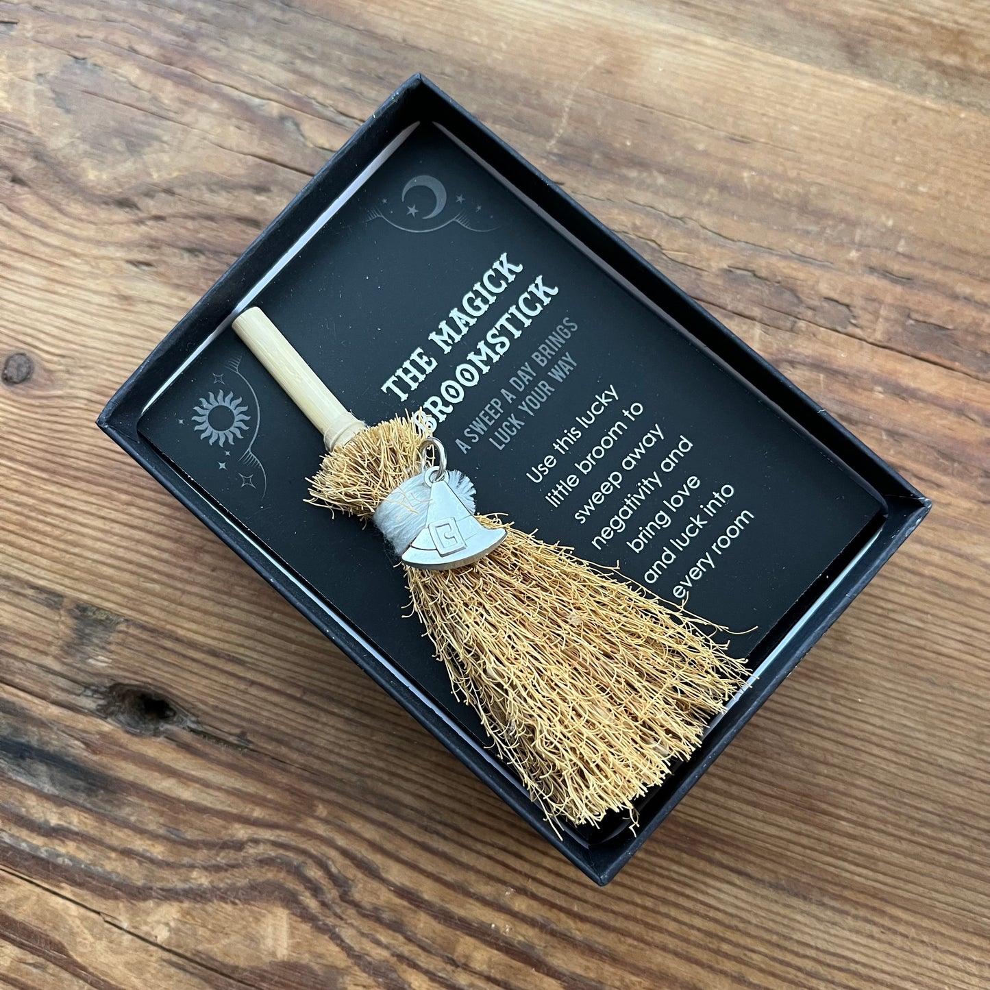 A small decorative broomstick gift set in a black presentation box on a wooden tabletop. The broom has a light wood handle, tan bristles, and a silver witch's hat charm