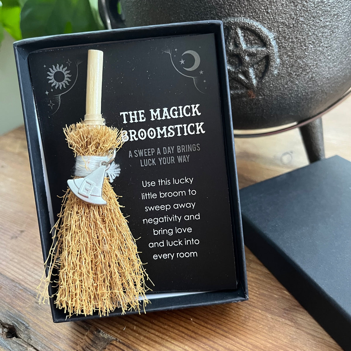 A small decorative broomstick gift set in a black presentation box on a wooden surface. The broom has natural straw bristles, a light wood handle, a silver witch's hat charm, and a black card with white text reading 'THE MAGIC BROOMSTICK' and additional blessing text.