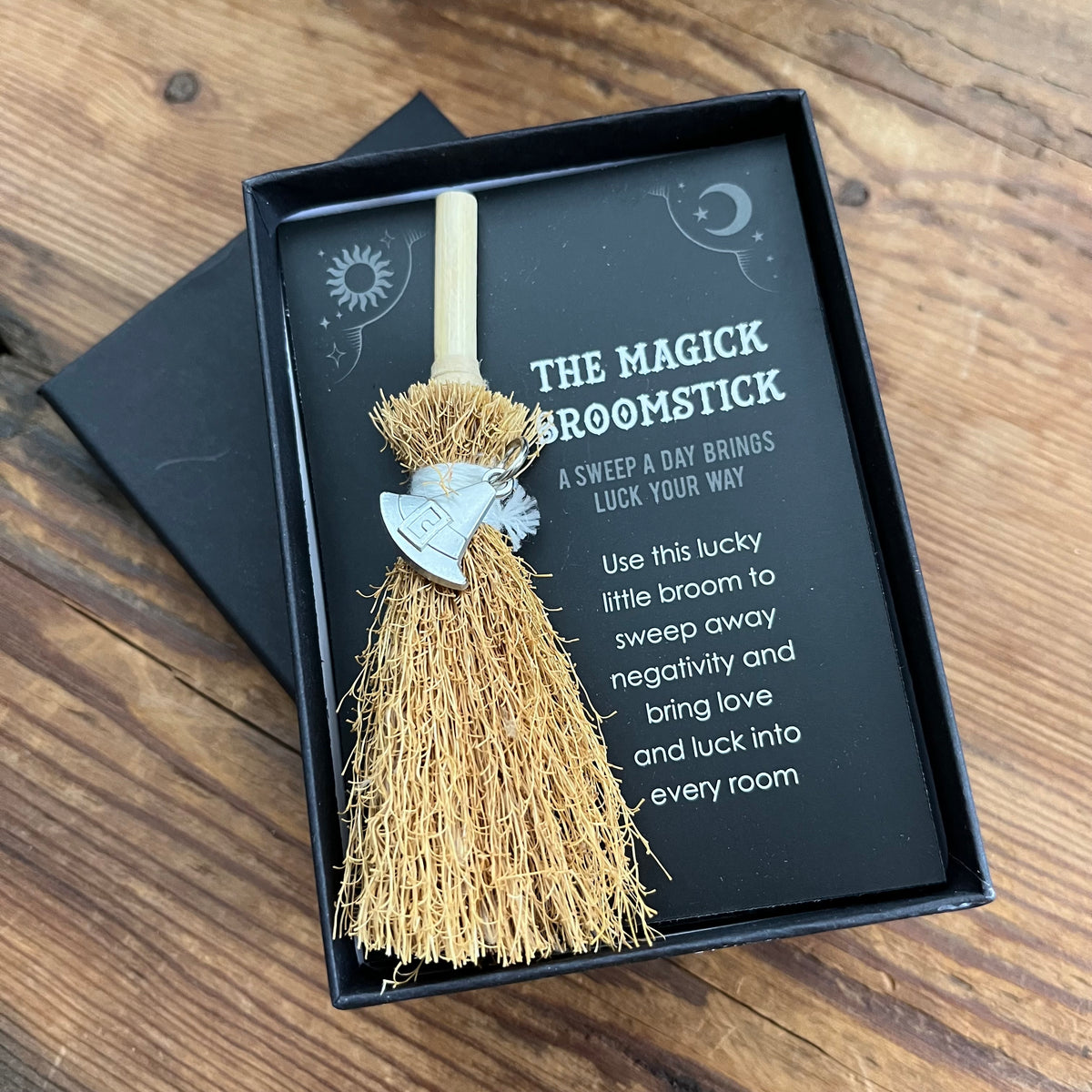 A packaged broomstick charm displayed in a black gift box on a wooden surface. The product is a small straw-colored broom with a light wood handle and a silver metal witch's hat charm, presented with printed card text reading 'THE MAGICK BROOMSTICK'.