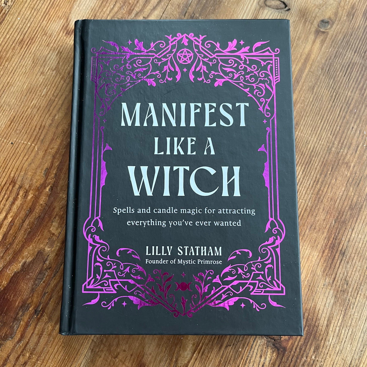 A hardcover book titled 'Manifest Like a Witch' by Lilly Statham, featuring a dark charcoal cover with ornate purple filigree borders and light blue lettering, placed on a wooden tabletop.