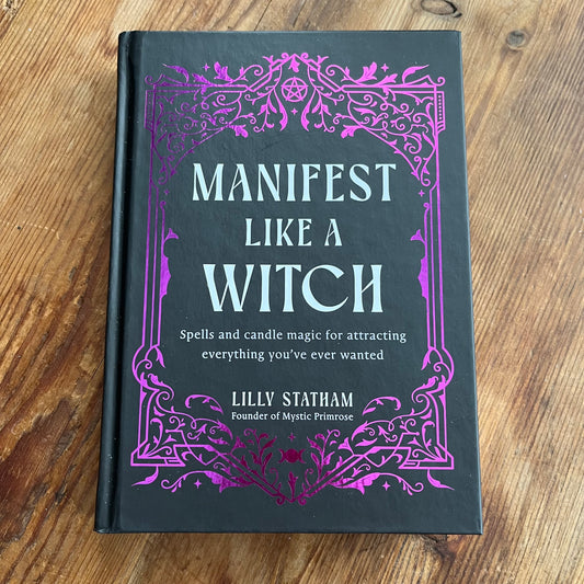 A hardcover book titled 'Manifest Like a Witch' by Lilly Statham, featuring a dark charcoal cover with ornate purple filigree borders and light blue lettering, placed on a wooden tabletop.