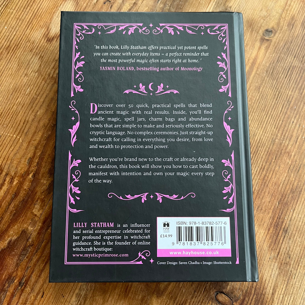 The back cover of the paperback book 'Lily Statham: Practical Witchcraft' by Lily Statham, featuring a black background with pink decorative borders and text, placed on a wooden surface.