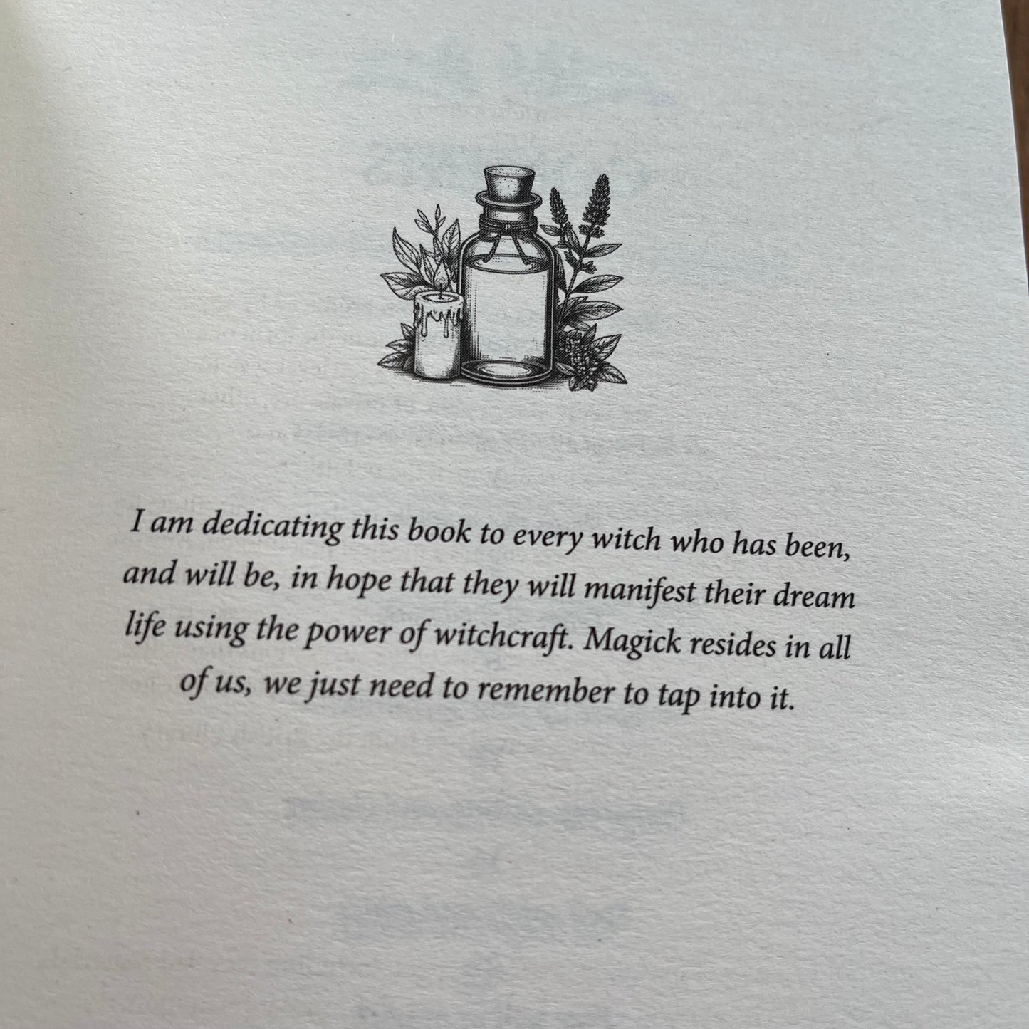 A close-up of a book's dedication page featuring a black ink illustration of a potion bottle and candle, with a handwritten-style dedication text below on aged off-white paper.